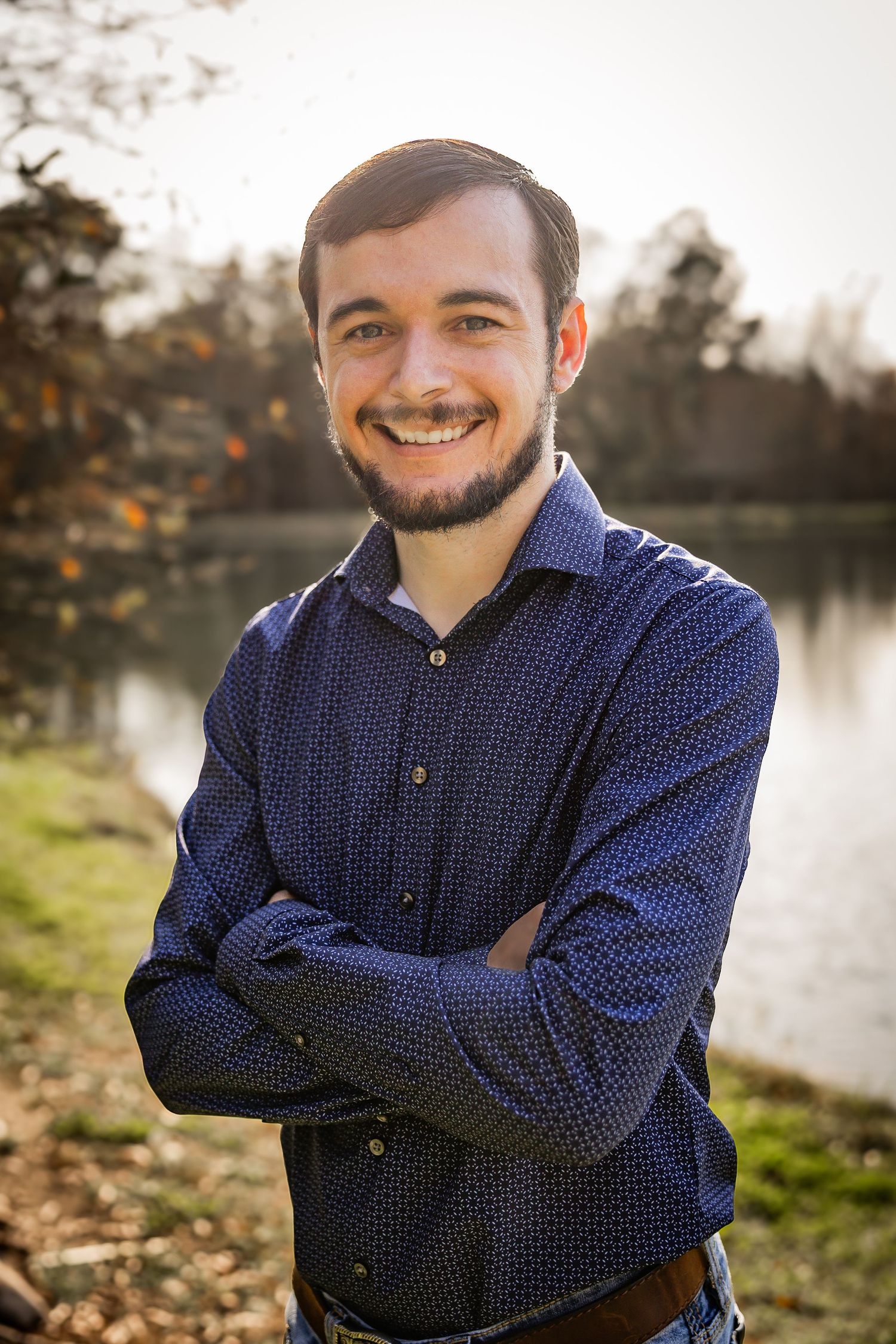 Confident young bearded man smiling with arms crossed outdoors near a sunlit autumn lake.