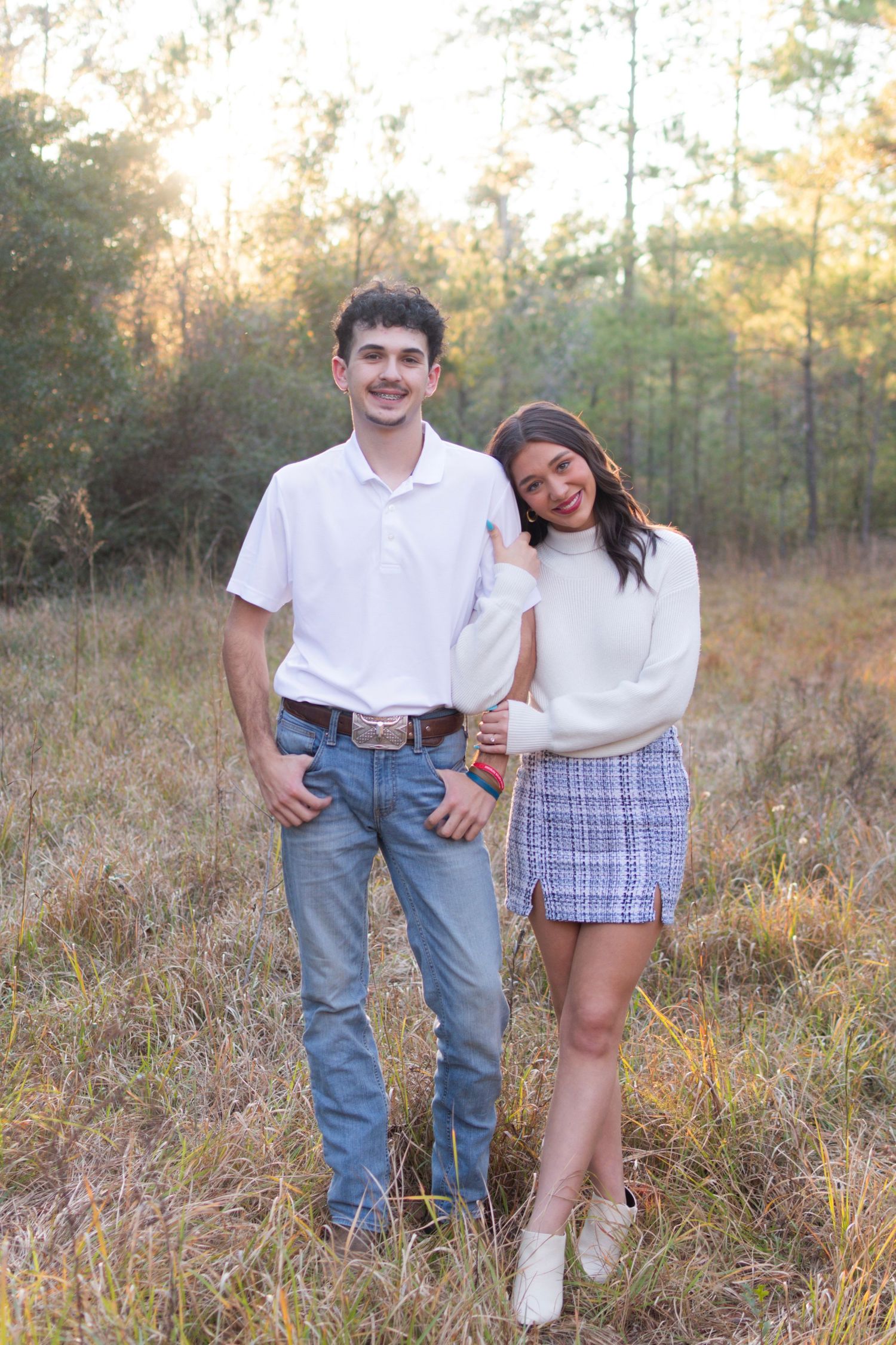 Young couple standing together in a golden field at sunset, man in white polo and woman in white sweater and plaid skirt.