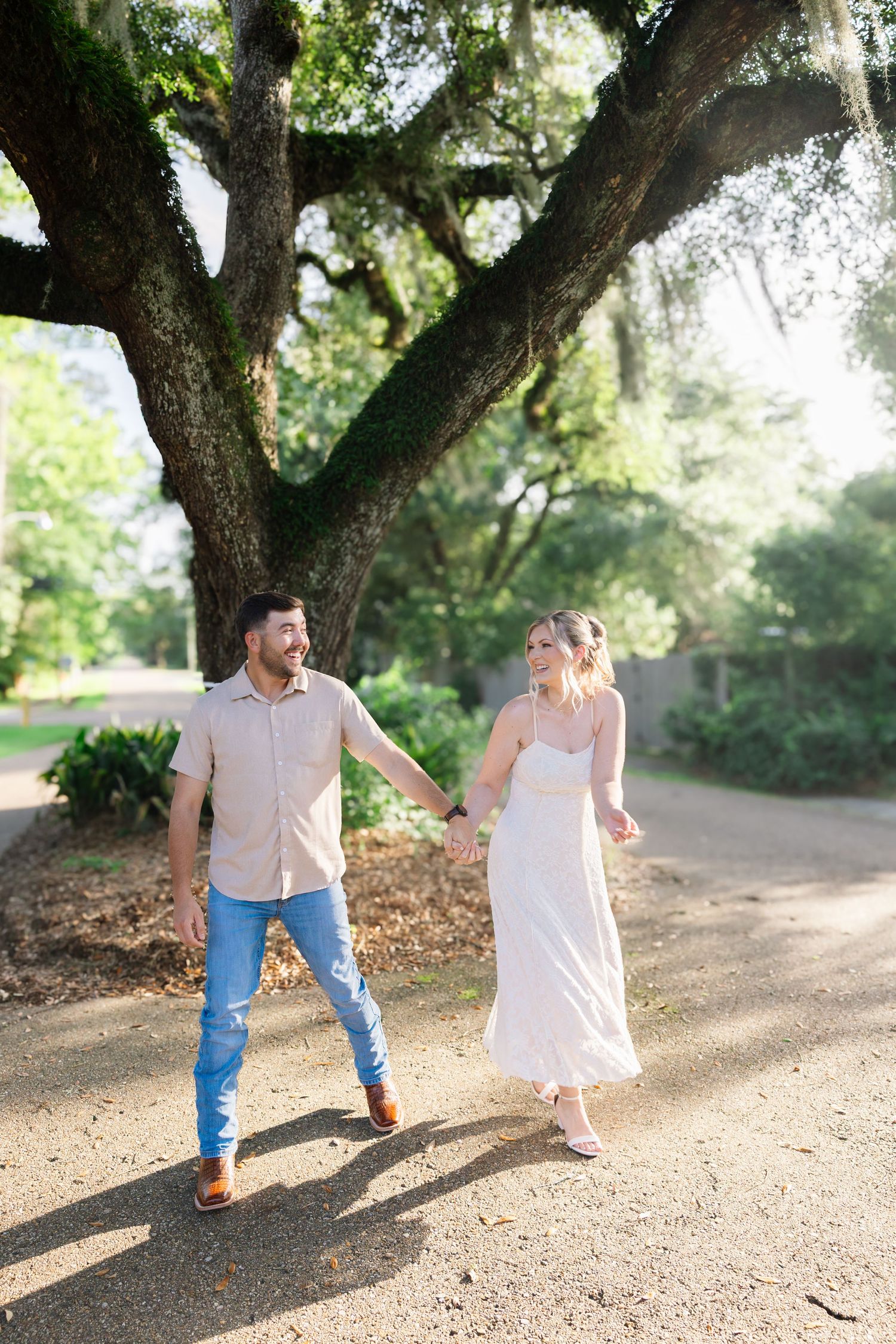 A happy couple holds hands and walks along a scenic gravel path beneath a large moss-covered oak tree.