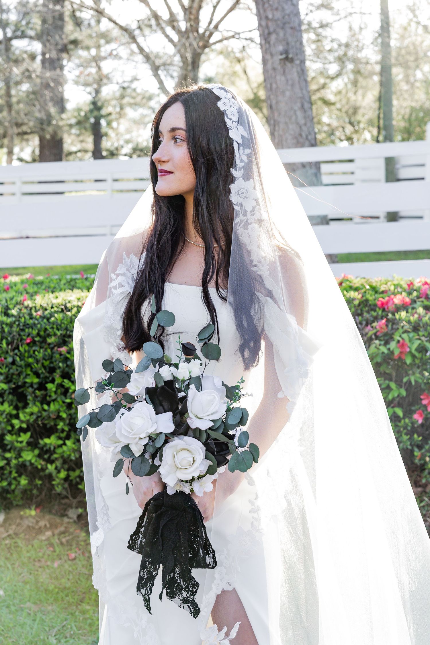 Beautiful bride in white gown holds a bouquet with black lace accents in a garden.