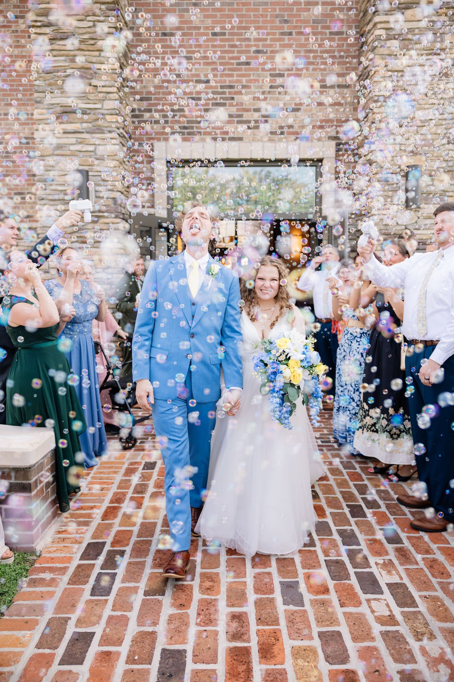 Bride and groom exit wedding ceremony through bubble send-off on brick pathway, surrounded by cheering guests.