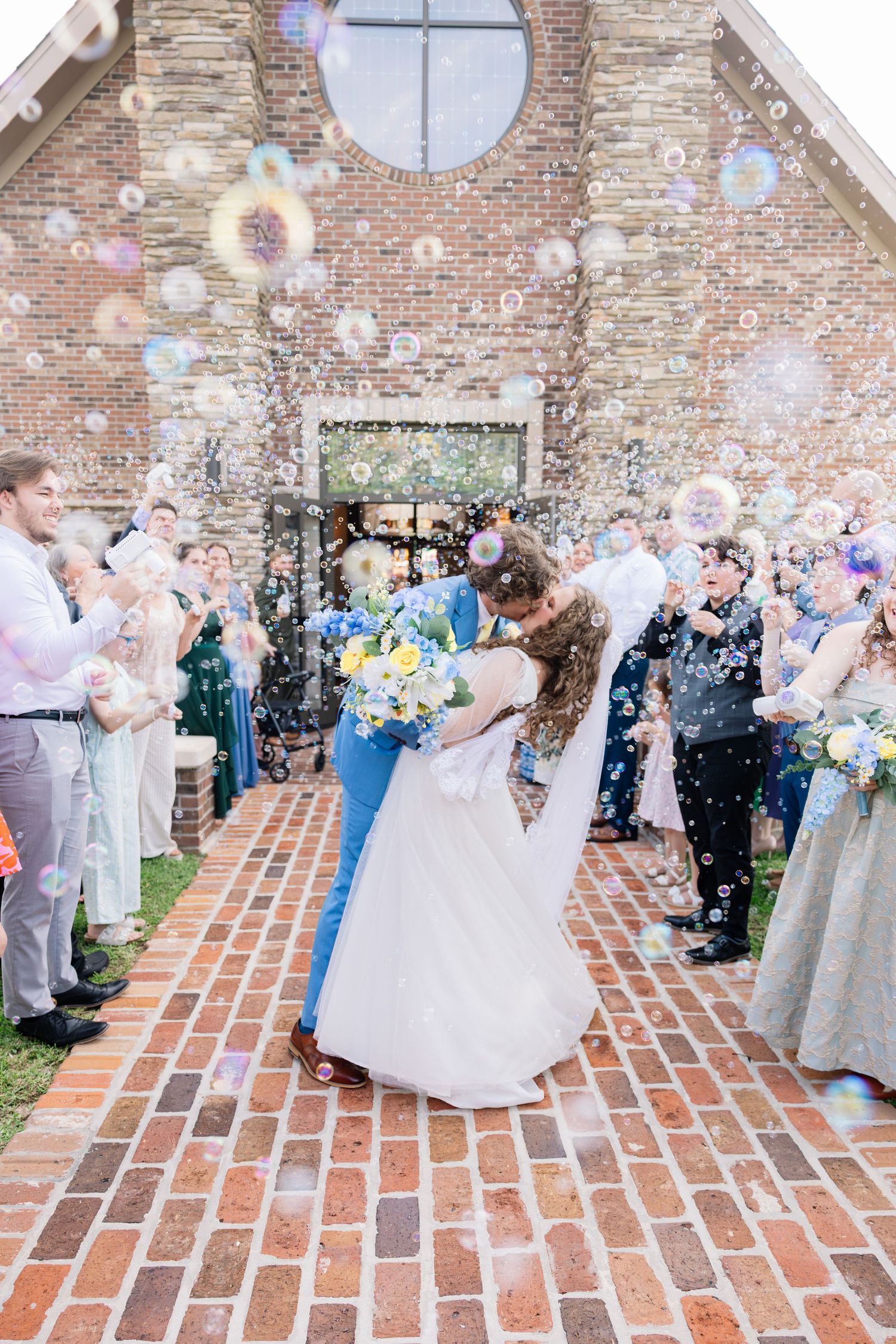 Bride and groom share a romantic dip kiss outside brick church surrounded by bubbles and cheering wedding guests.