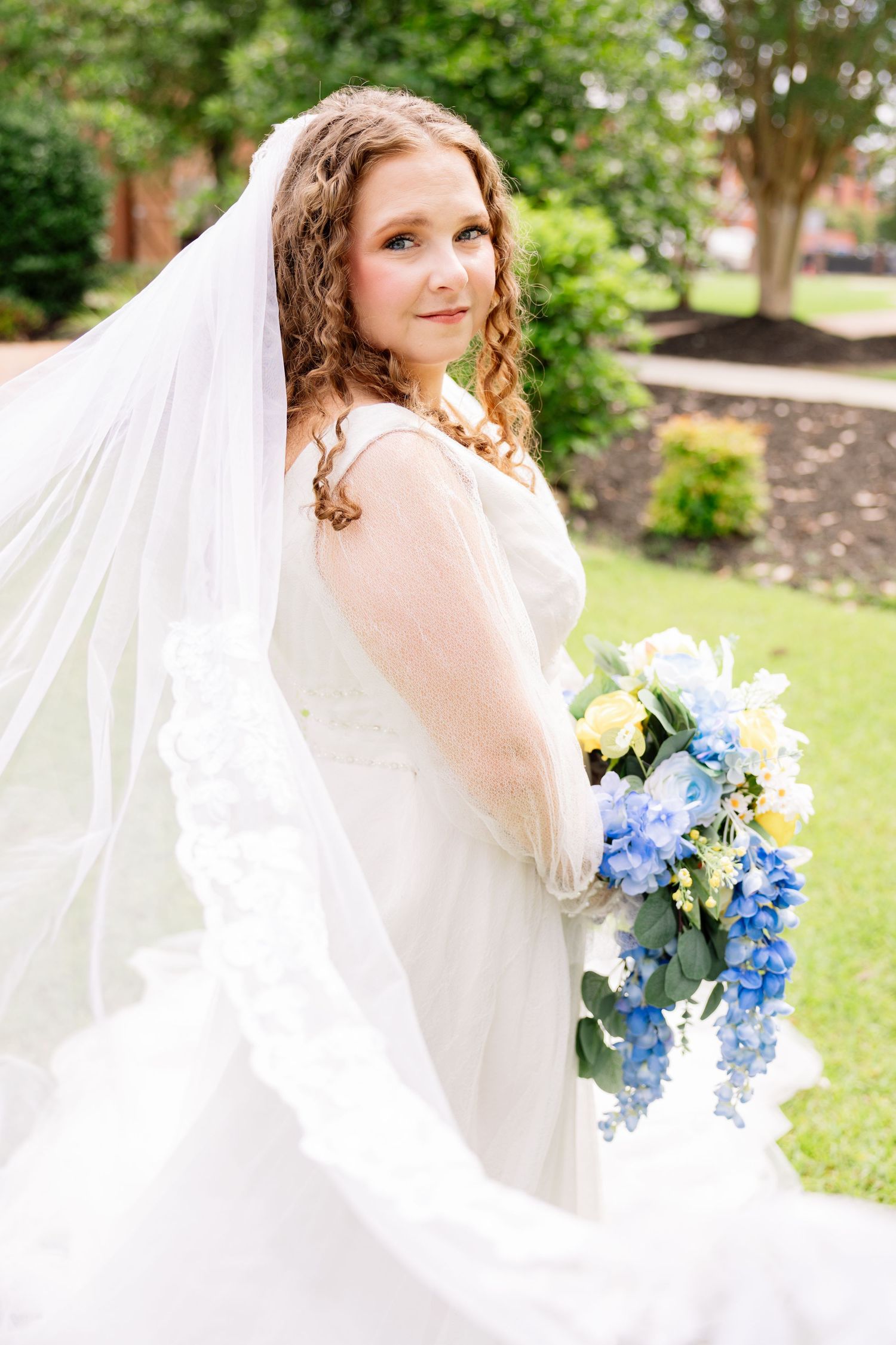 Bride in white gown and flowing veil holds blue and yellow bouquet outdoors on green lawn near brick building.