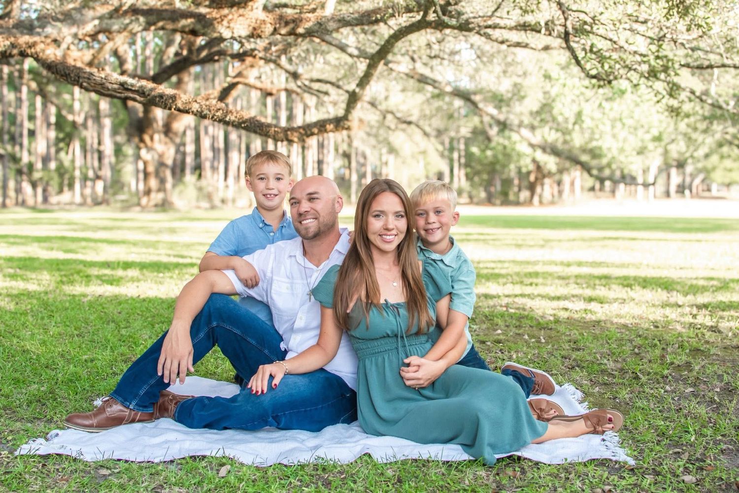 Happy family of four sits on blanket under oak trees in lush green park for outdoor portrait.