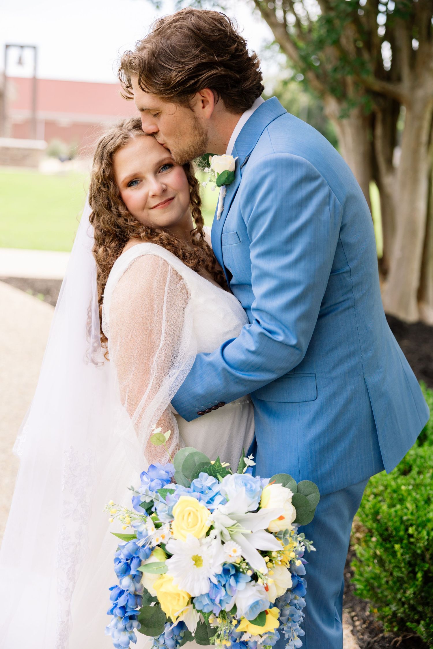 Groom in blue suit kisses bride with curly hair holding blue and yellow bouquet outdoors.