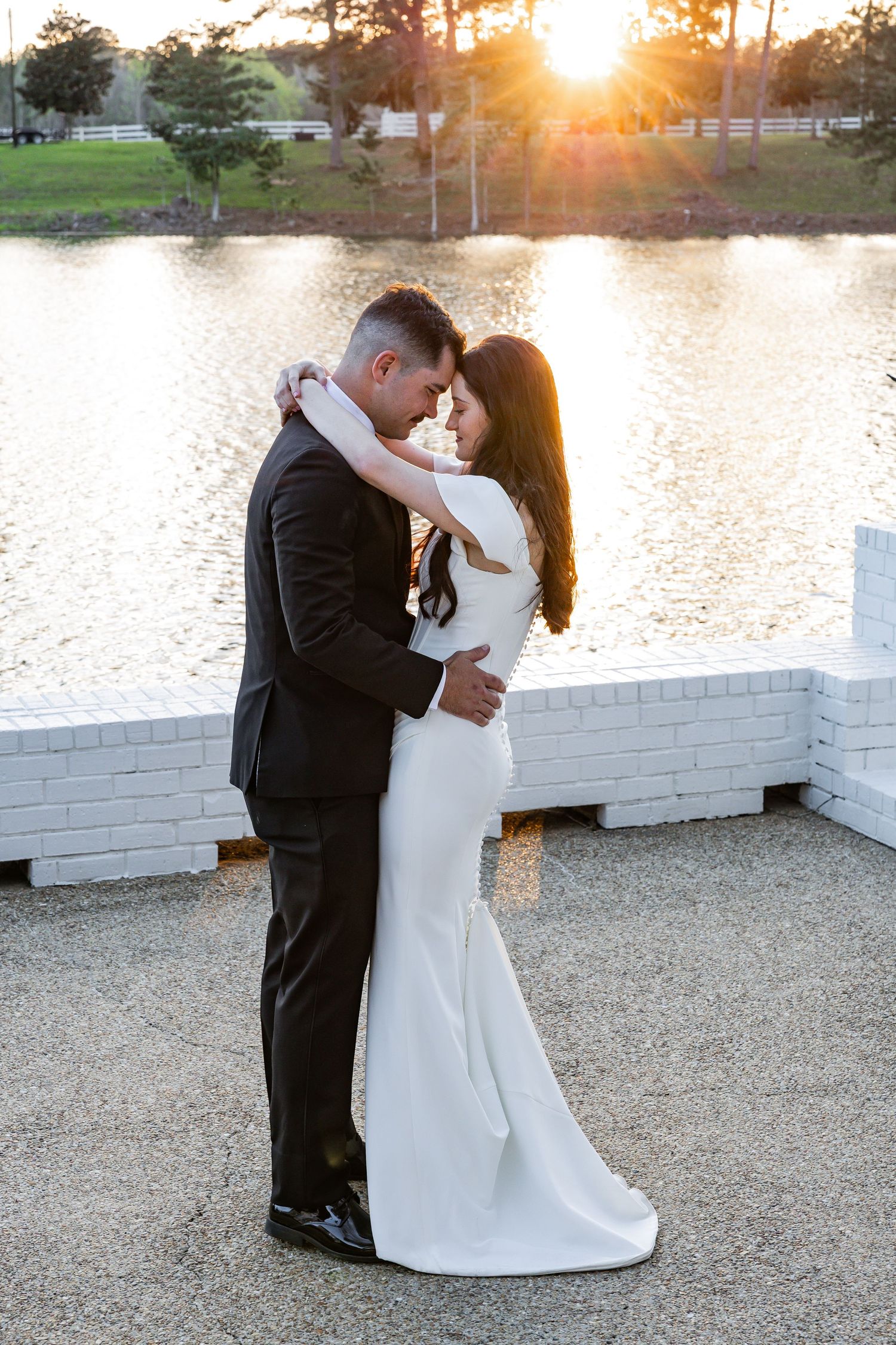 Bride and groom embracing forehead-to-forehead by a sunlit lake in a romantic sunset portrait.