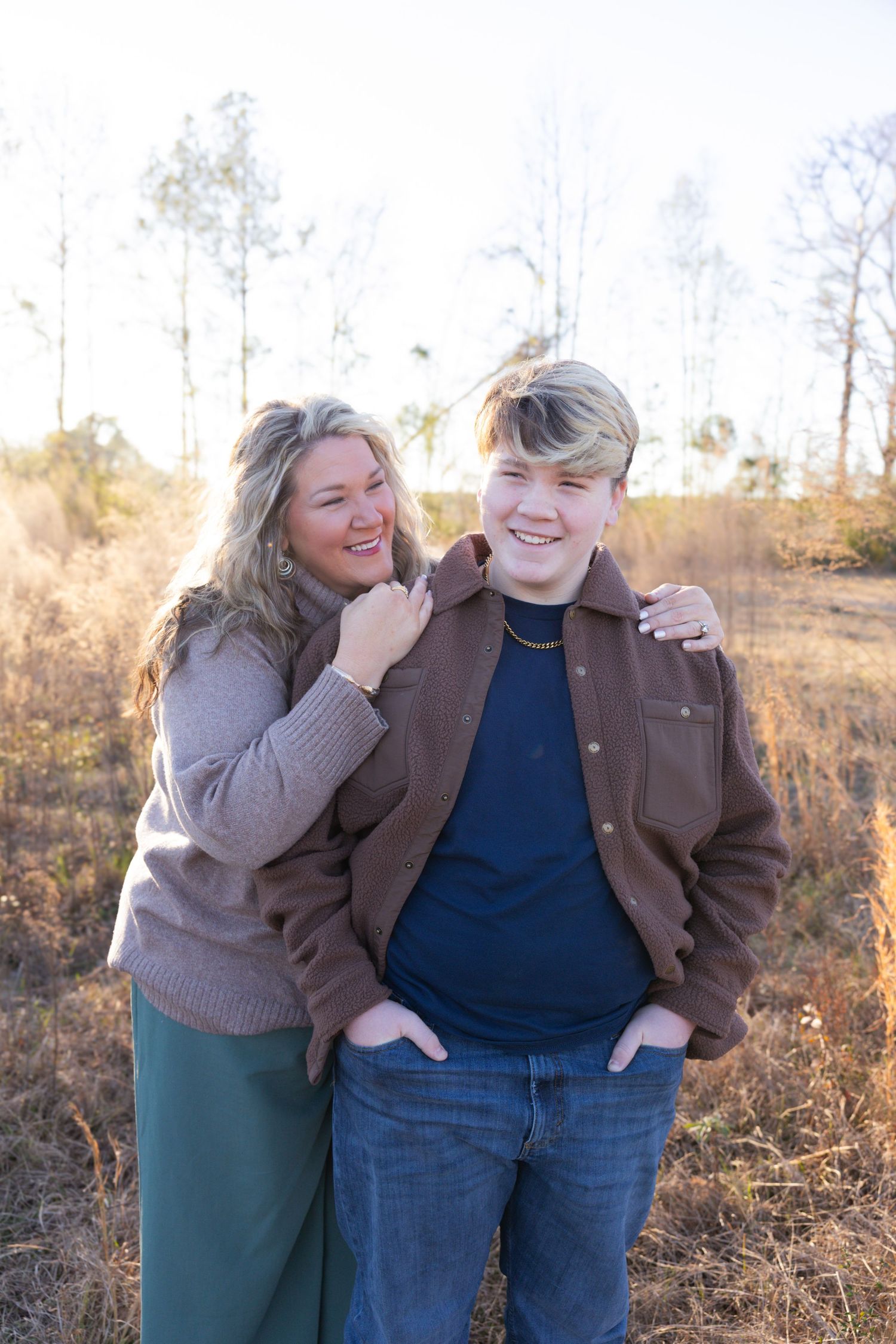 Mother and teenage son smiling together outdoors in a golden field during warm sunset light.