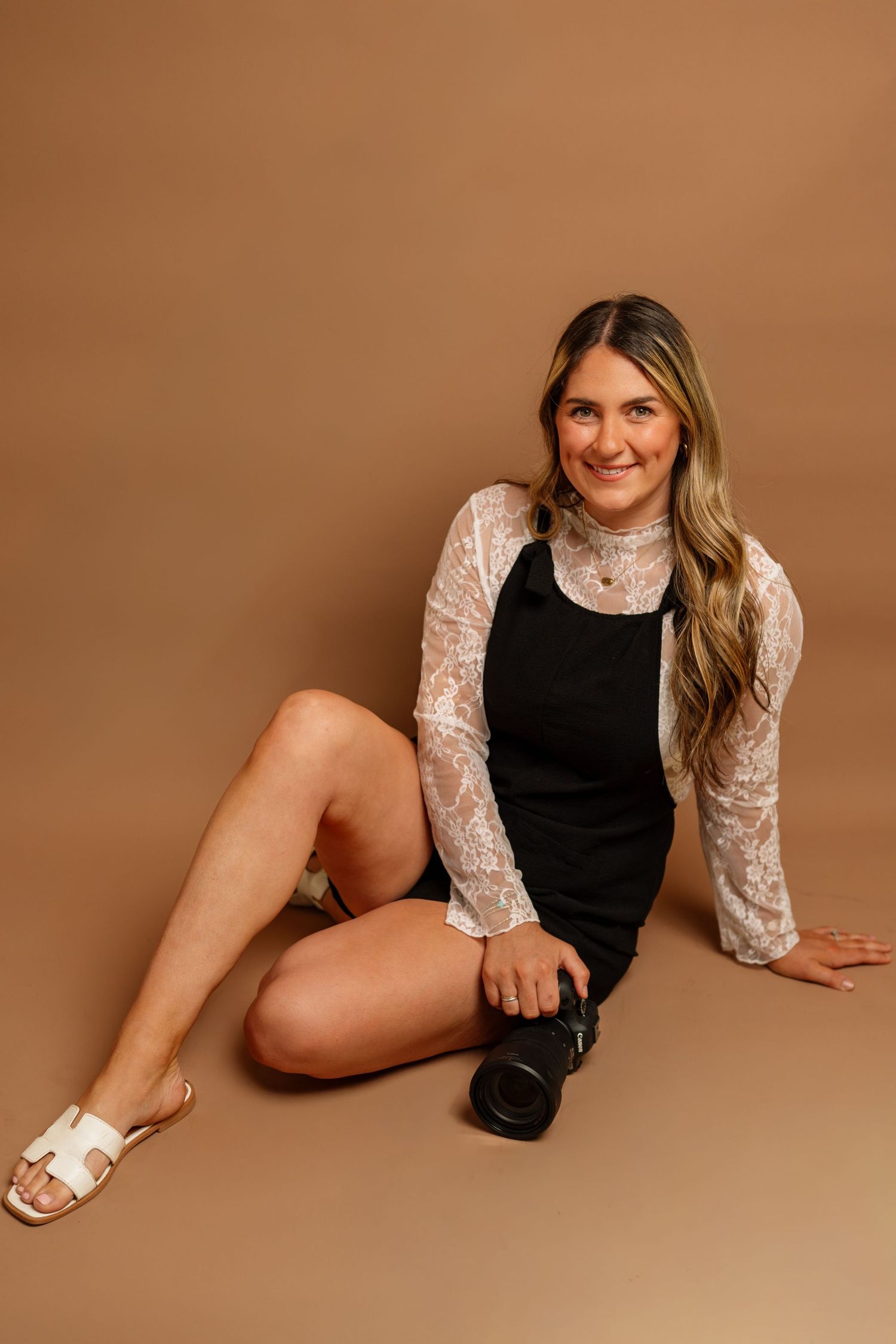 Young woman in black dress and lace cardigan sitting on studio floor holding a camera, smiling warmly.