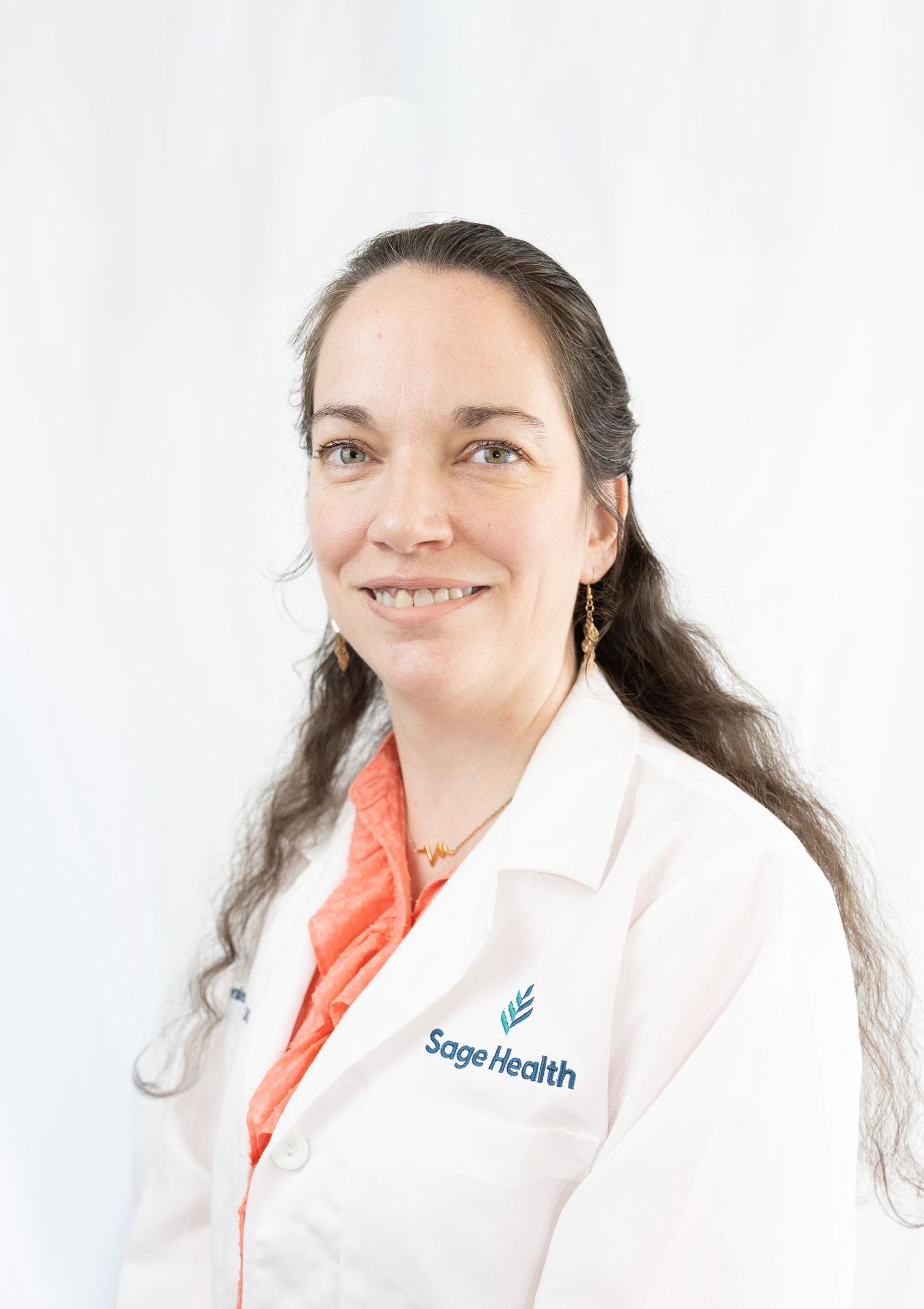 Female healthcare professional in white Sage Health lab coat smiling against white background.