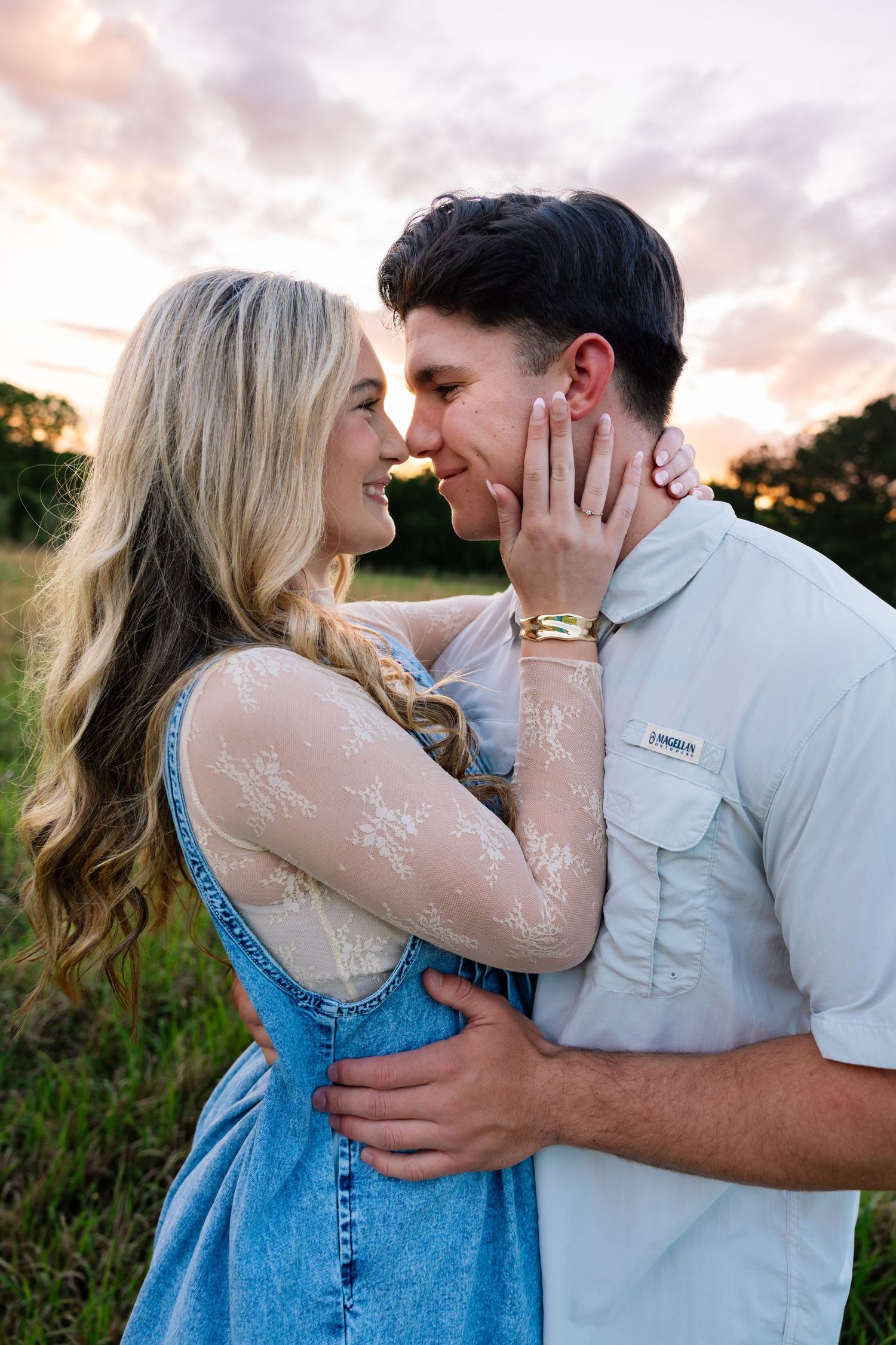 Couple sharing a romantic almost-kiss at golden hour in a green field, woman in denim dress touching mans face.