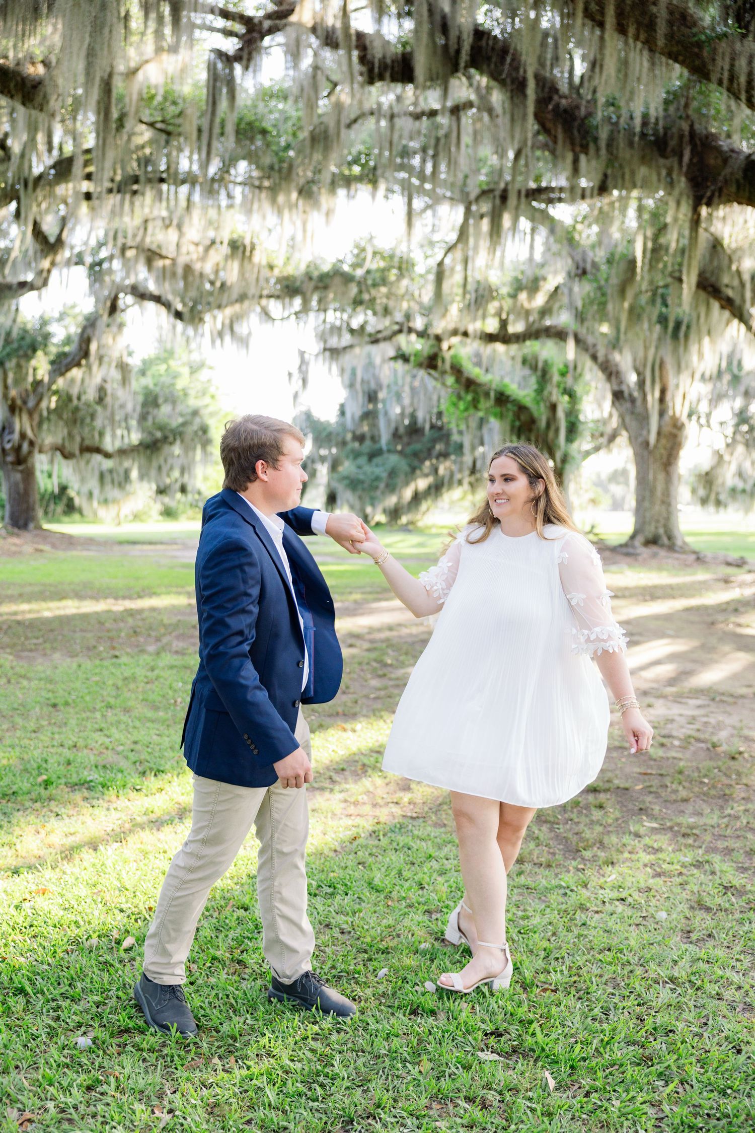 A couple dances under moss-draped oak trees, woman in white dress, man in navy blazer, on lush green grass.