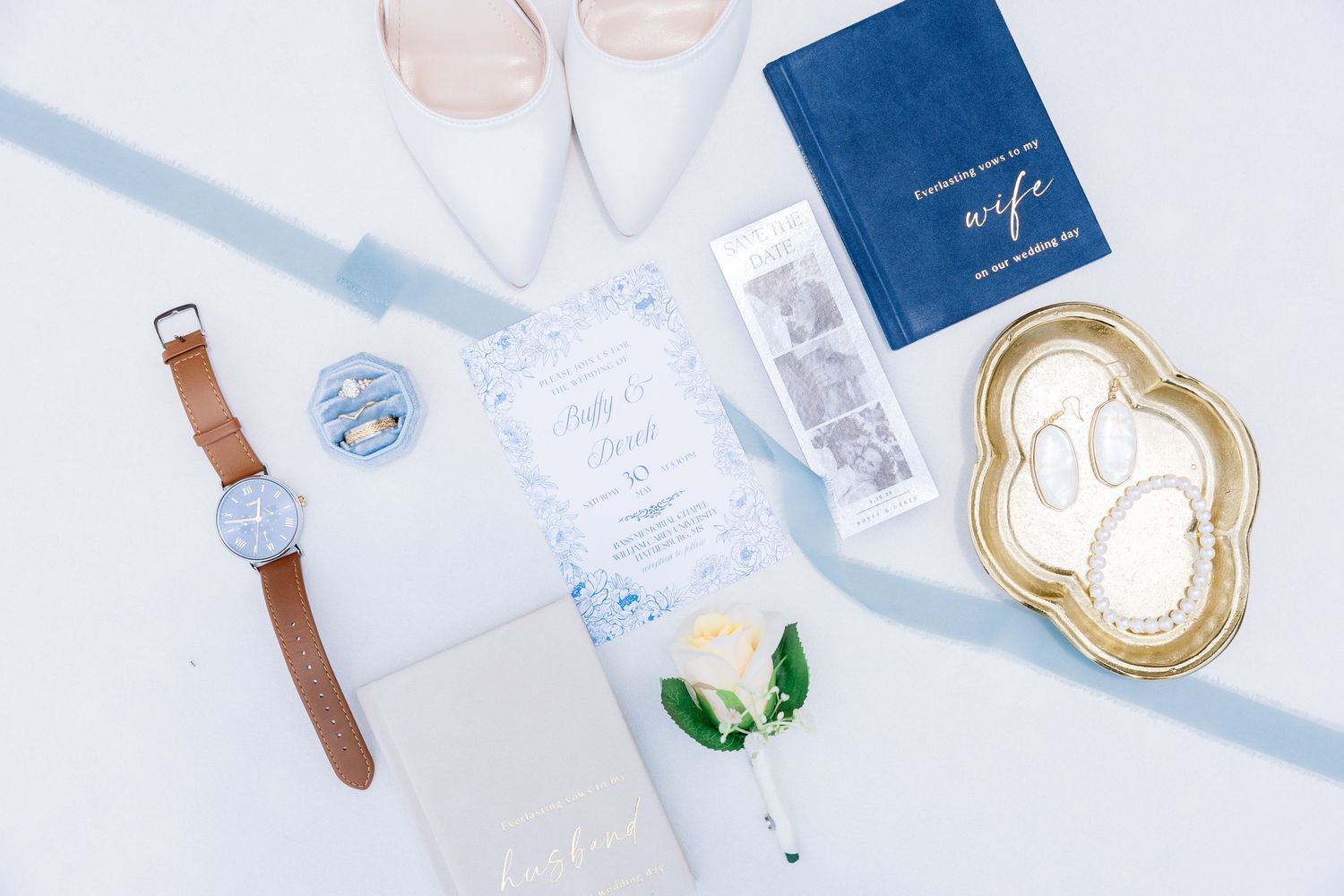 Elegant wedding flat lay with blue invitation, white heels, passport, watch, rings, and boutonniere on light blue background.