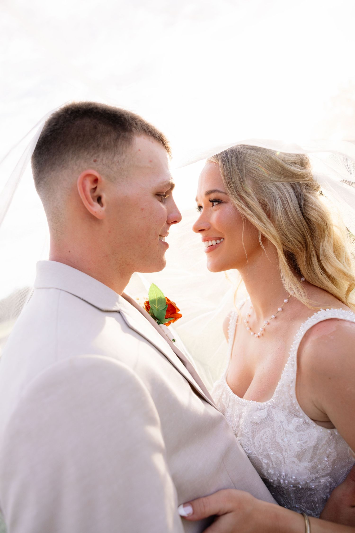 Bride and groom gazing at each other under a veil, groom in beige suit with boutonniere, bride in lace gown.