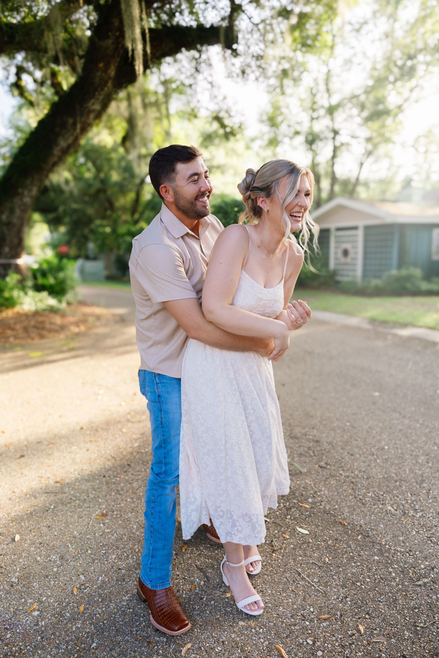 A joyful couple embraces and laughs together on a sunlit gravel path surrounded by lush green trees.