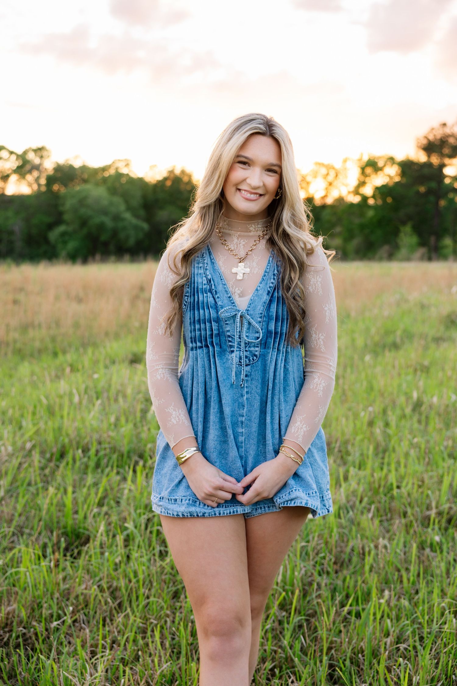 Young blonde woman in denim romper and cross necklace smiling in a grassy field at golden hour sunset.