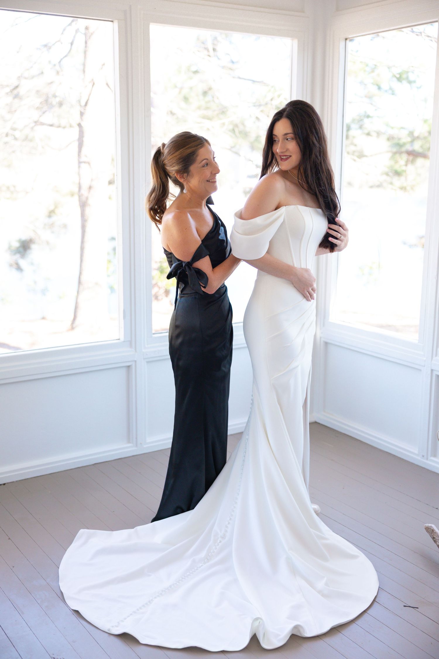 Bridesmaid in black dress assisting smiling bride in elegant white off-shoulder wedding gown.