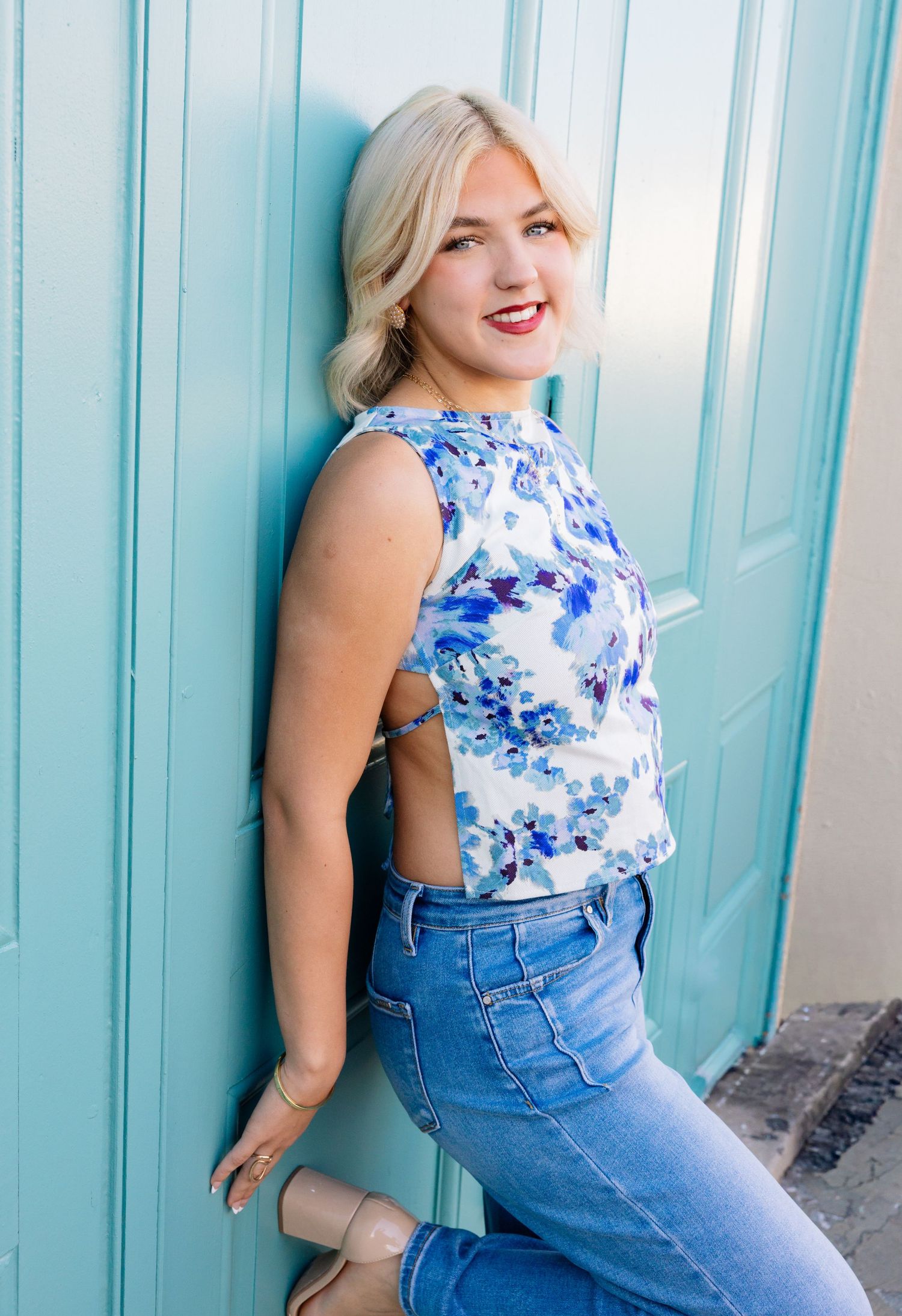 Young blonde woman in floral blue top and jeans leaning against a turquoise door, smiling over her shoulder.