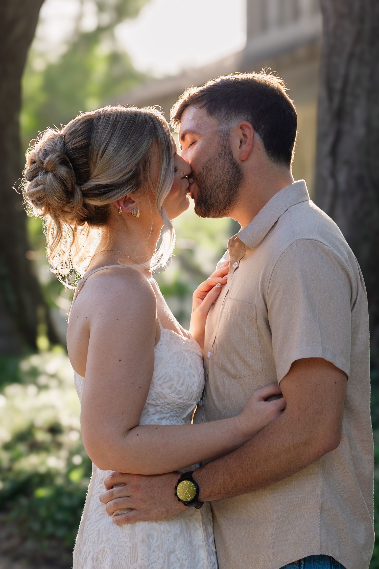 A couple shares a romantic kiss outdoors, the woman in a white dress with an updo, the man in a beige shirt, bathed in golden light.