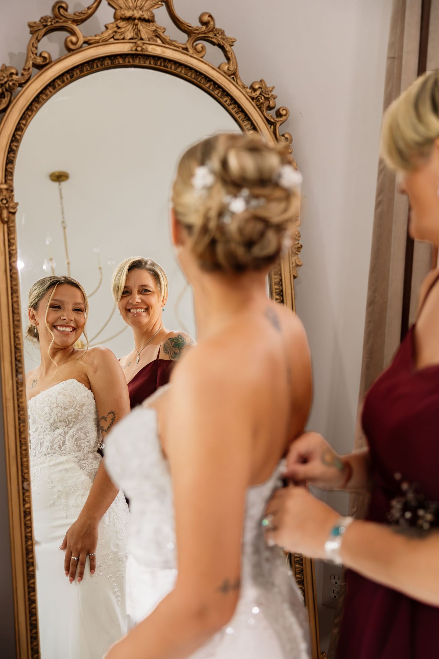 Bride in white lace gown getting dressed with bridesmaid in burgundy, reflected in ornate gold mirror.