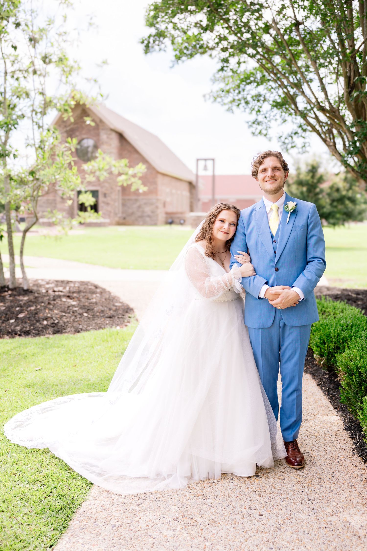 Bride in white gown and groom in blue suit pose together on a garden path outside a brick chapel.