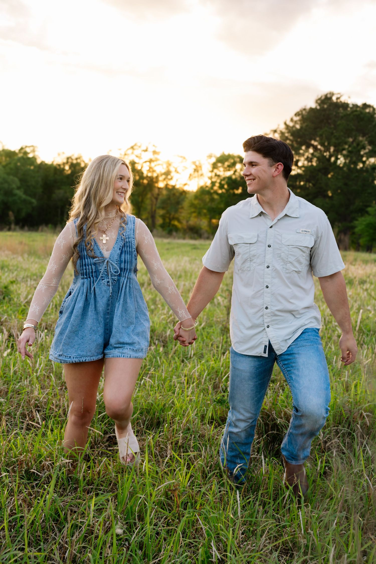 Happy couple holding hands walking through tall grass field at sunset, woman in blue denim romper and man in gray shirt.