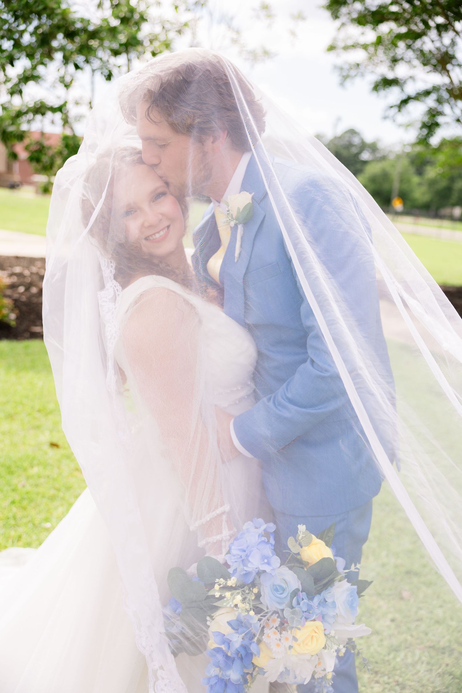 Bride and groom share a romantic kiss under a flowing white veil, bride holding blue and yellow bouquet outdoors.