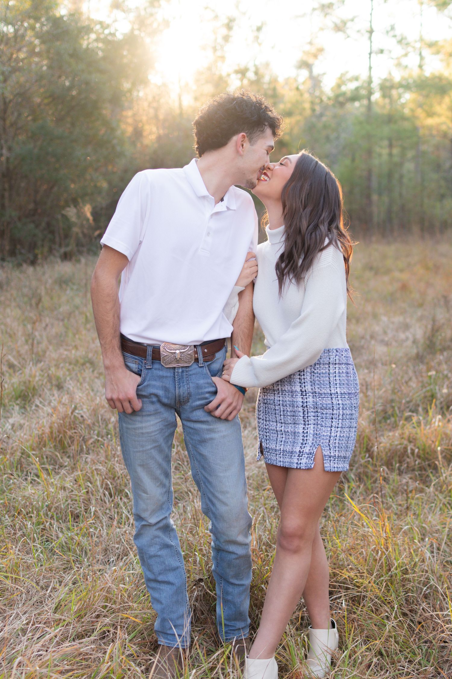 Young couple sharing a sweet kiss in a sunlit meadow, man in white polo jeans and woman in white sweater plaid skirt.
