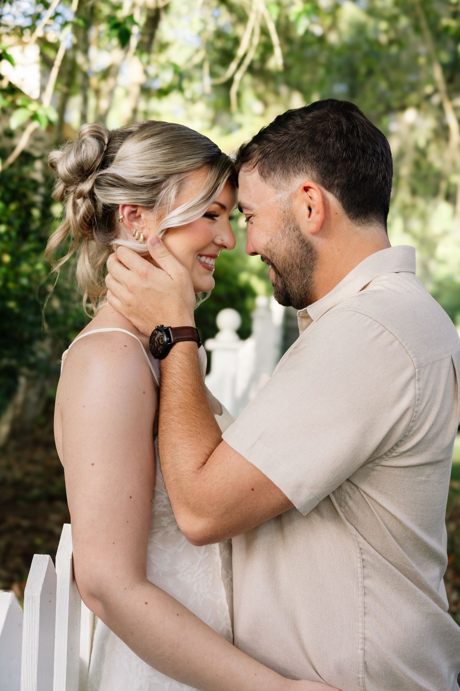A romantic couple shares an intimate moment, nearly touching foreheads, surrounded by lush green foliage.