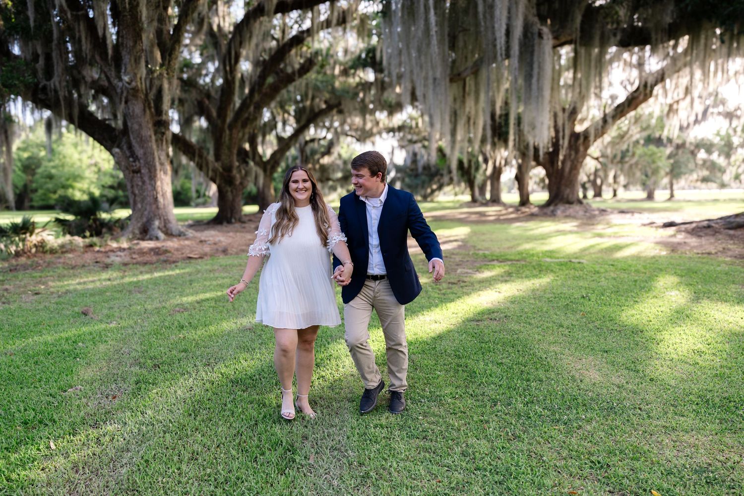 Happy couple laughing and walking through lush green park under canopy of moss-covered oaks.