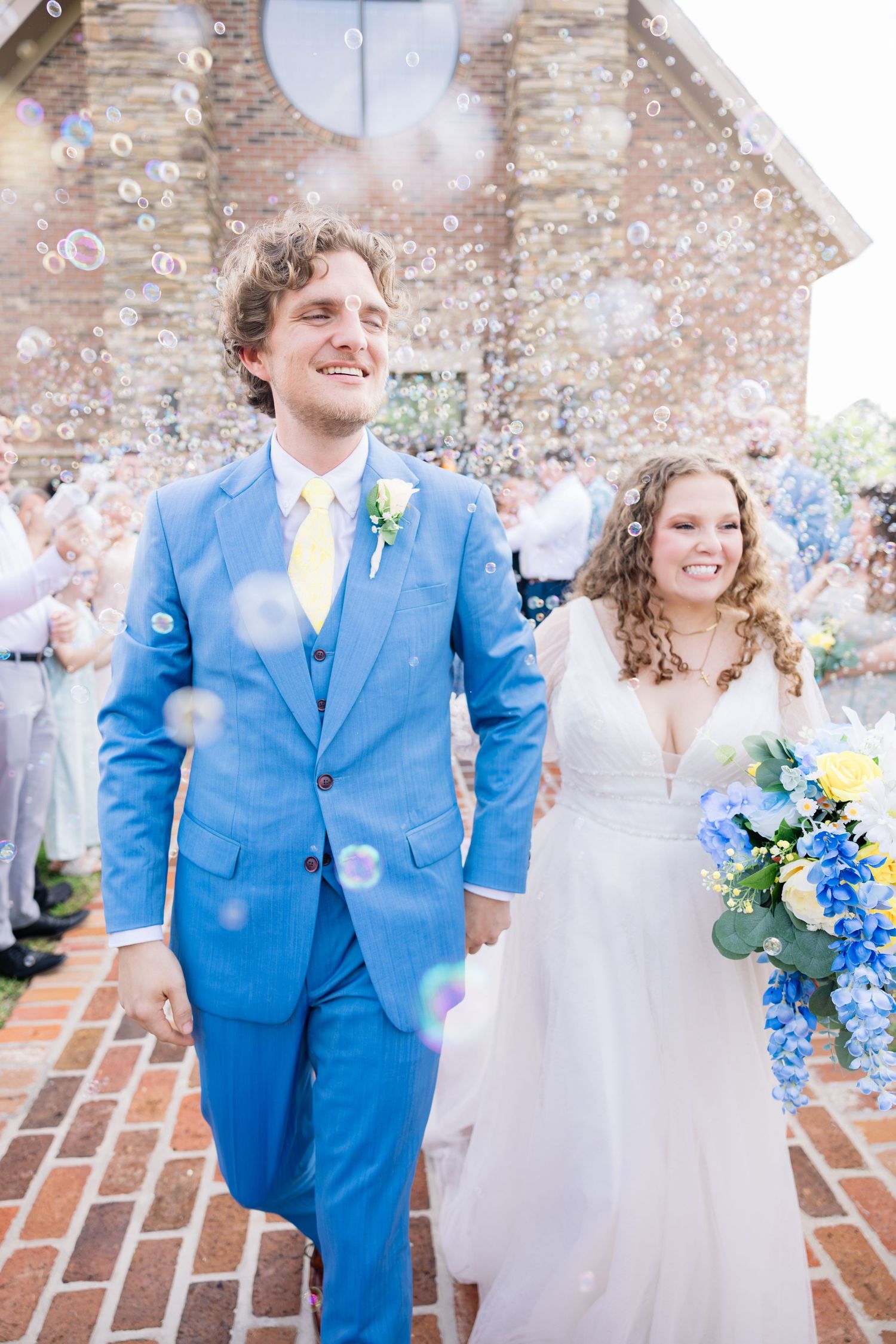 Newlywed couple exits church ceremony surrounded by bubbles, groom in blue suit, bride in white gown holding blue and yellow bouquet.