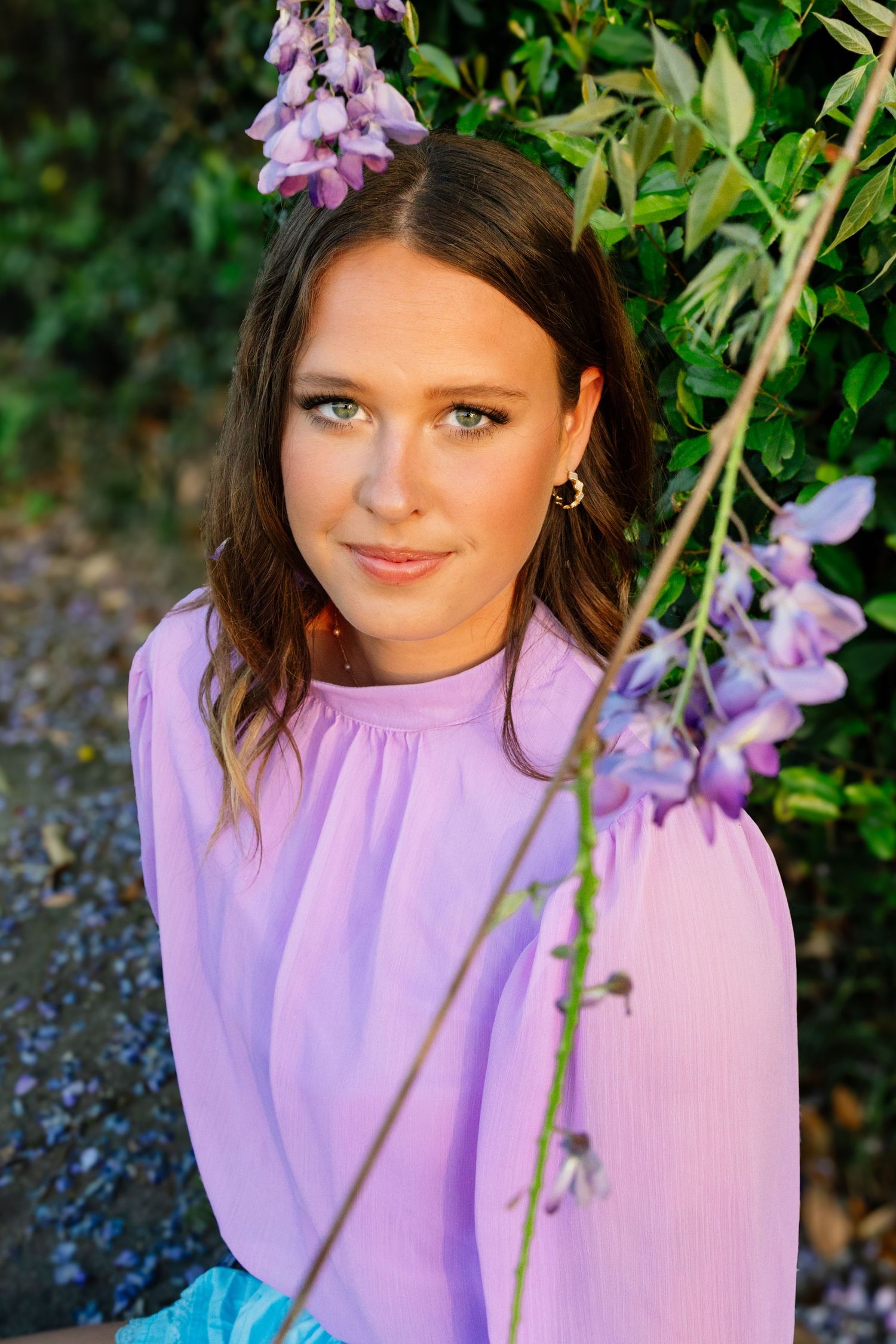 Young brunette woman in lavender blouse posing near purple wisteria blooms outdoors.