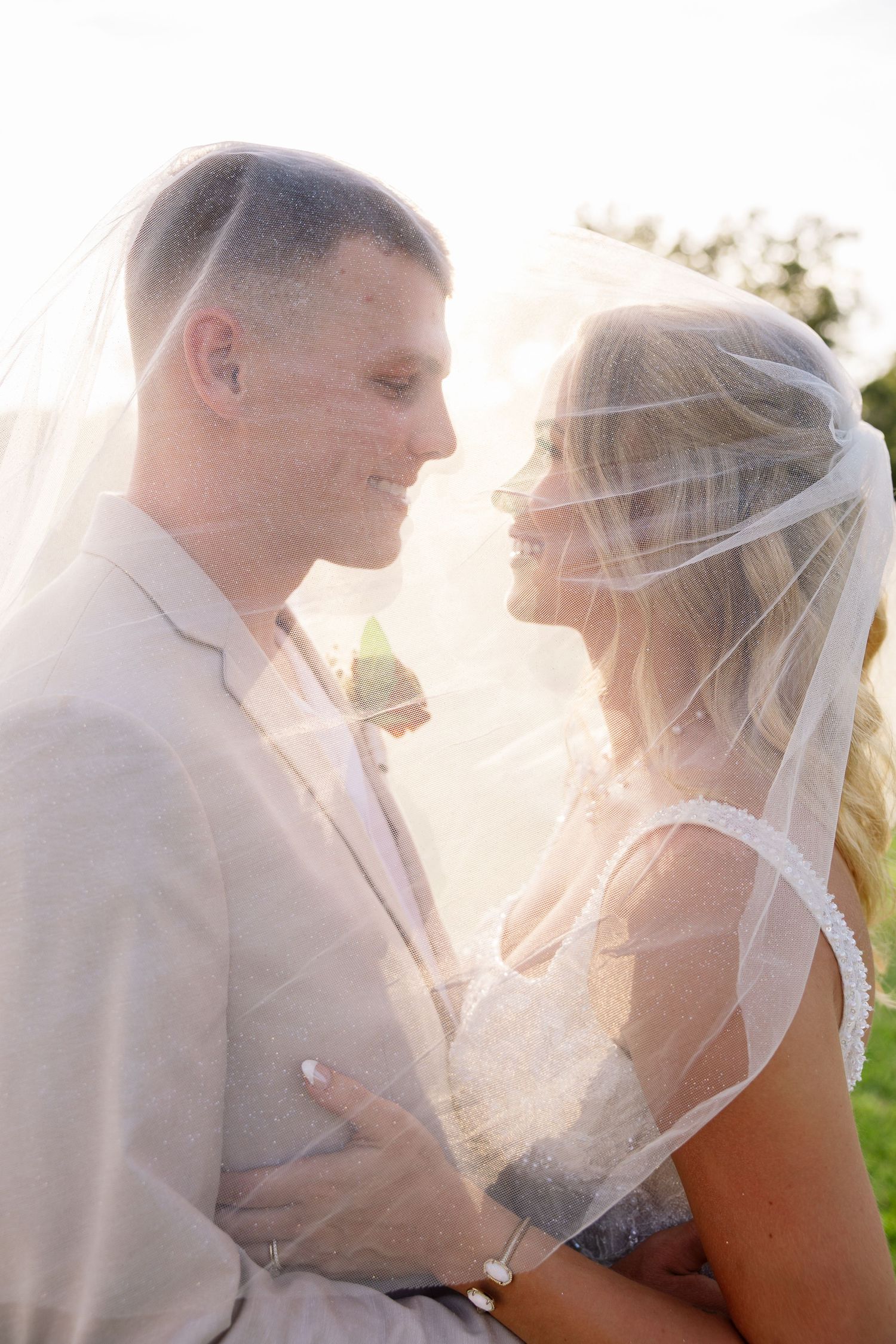 Bride and groom share a romantic moment under a flowing veil, bathed in soft golden sunlight outdoors.