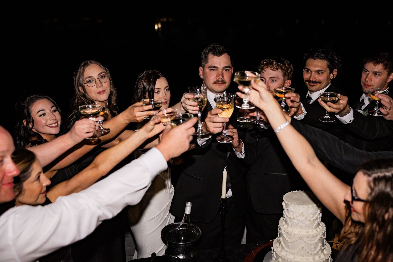 Wedding guests raising champagne glasses in a celebratory toast near a white tiered cake.