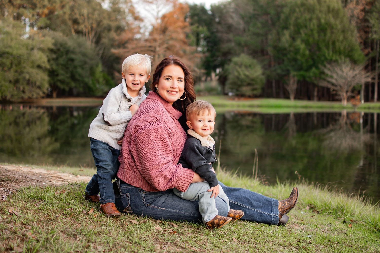 Happy mother sitting on grass by a lake with her two young sons in autumn setting.