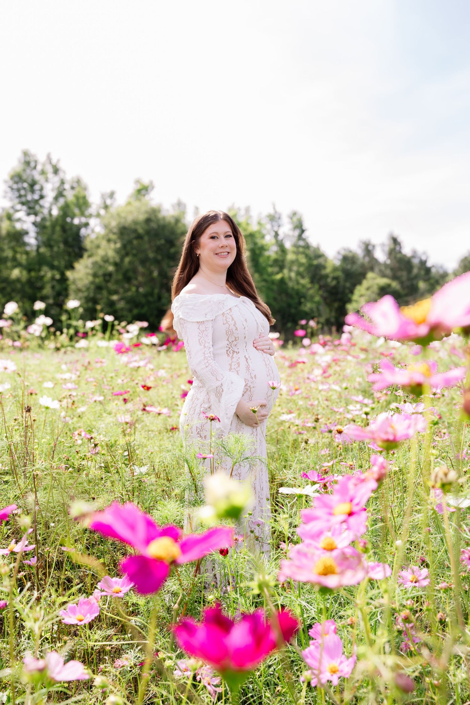 Pregnant woman in white lace dress standing in a blooming pink cosmos flower field with trees in the background.