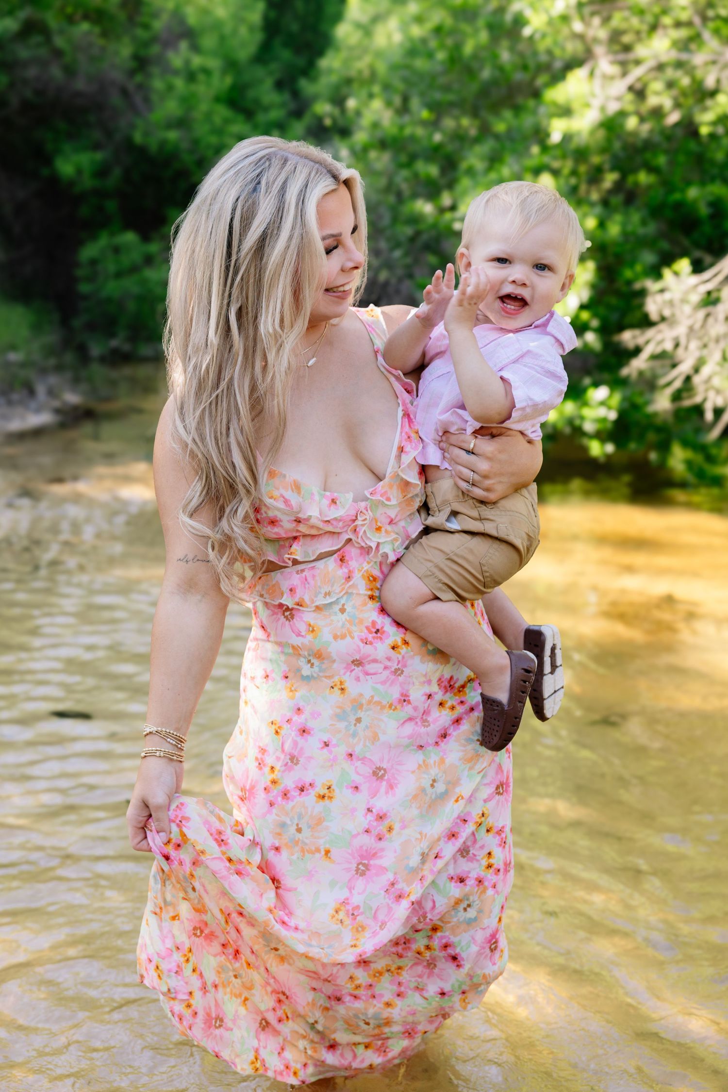 Blonde woman in pink floral maxi dress holding a cheerful baby while standing in a creek.