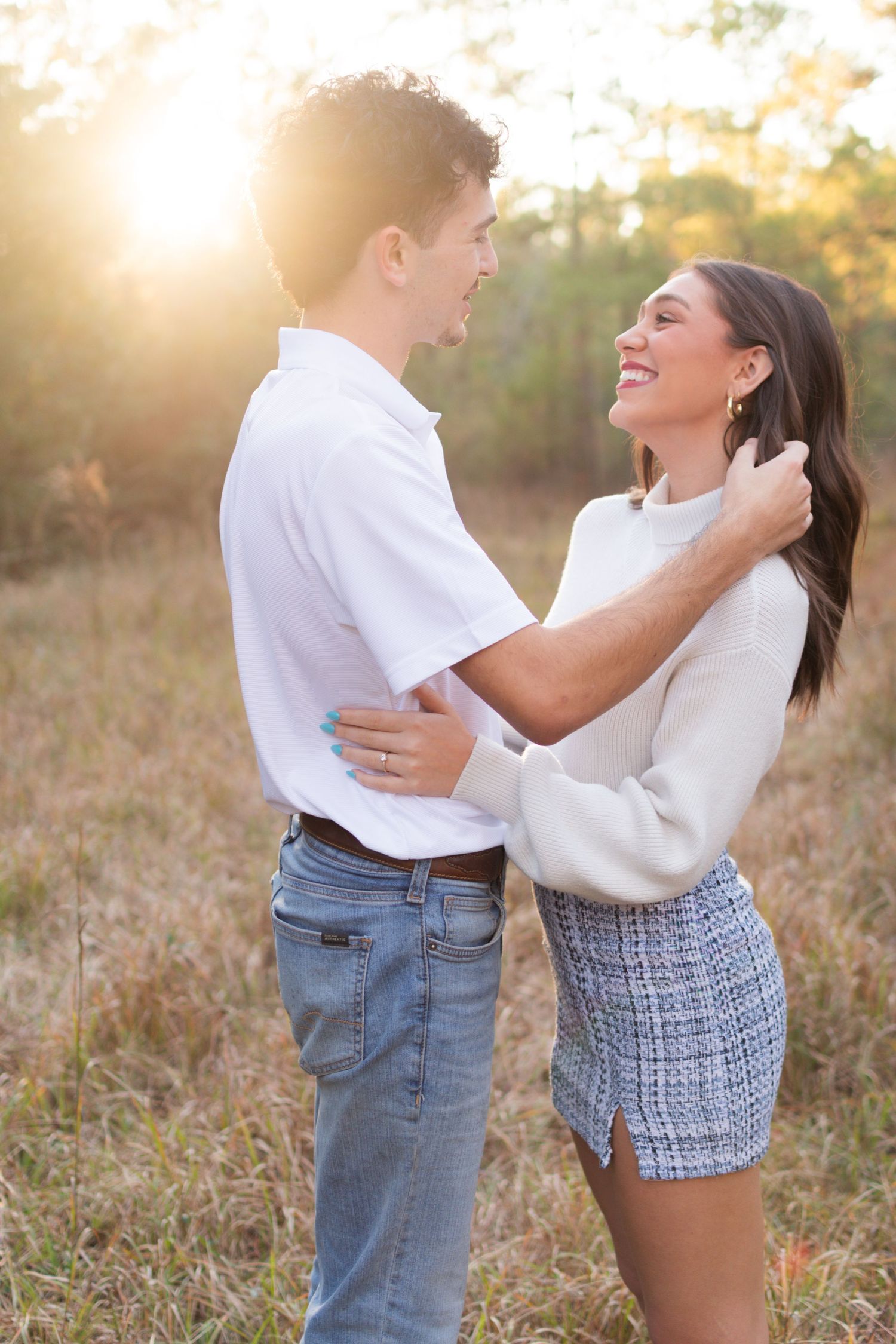 Joyful couple laughing and embracing in a golden sunlit field, man in white shirt and jeans, woman in white sweater.