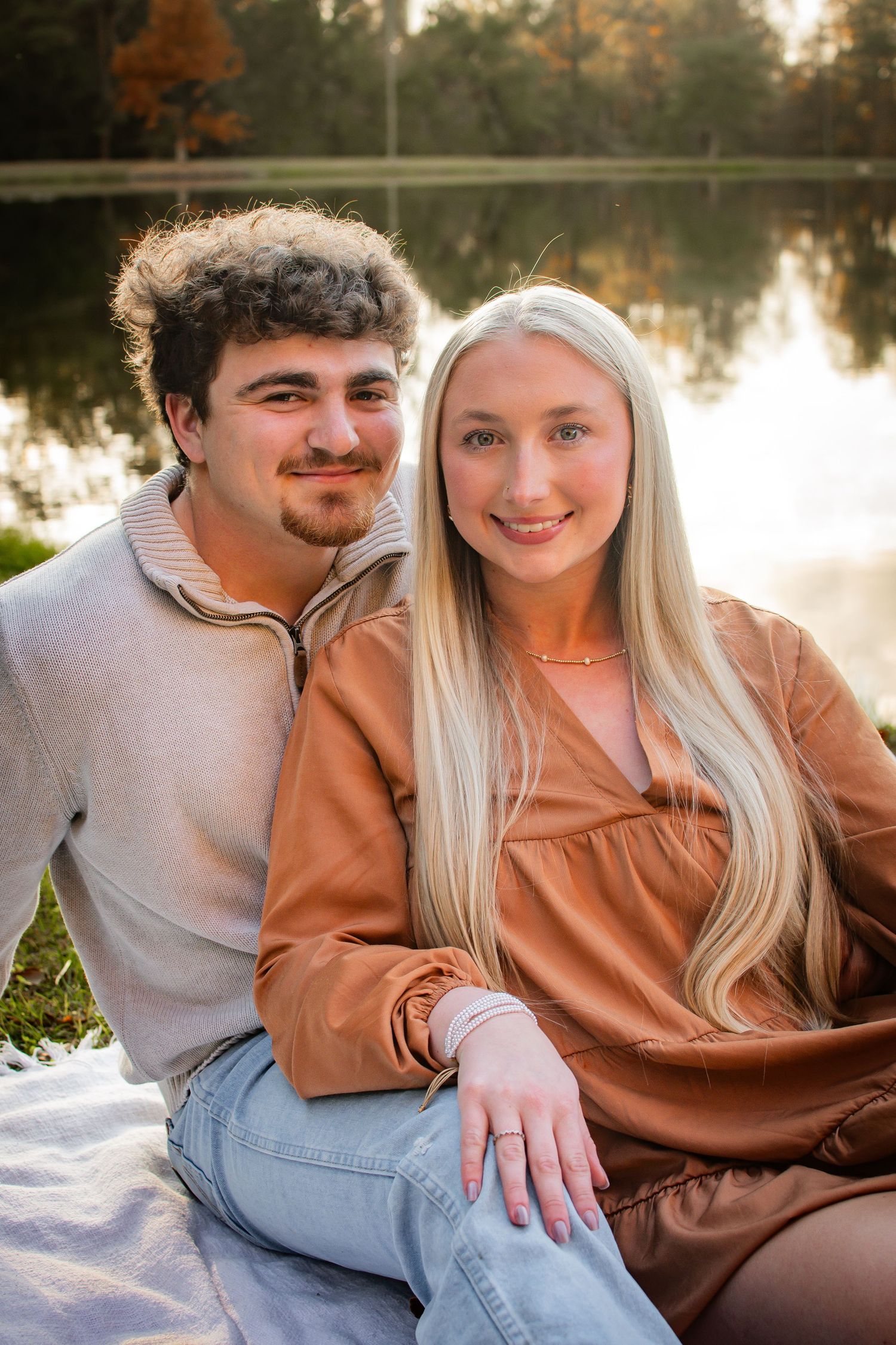 Smiling young couple sitting by a lake in autumn, woman in brown dress with engagement ring visible on her hand.