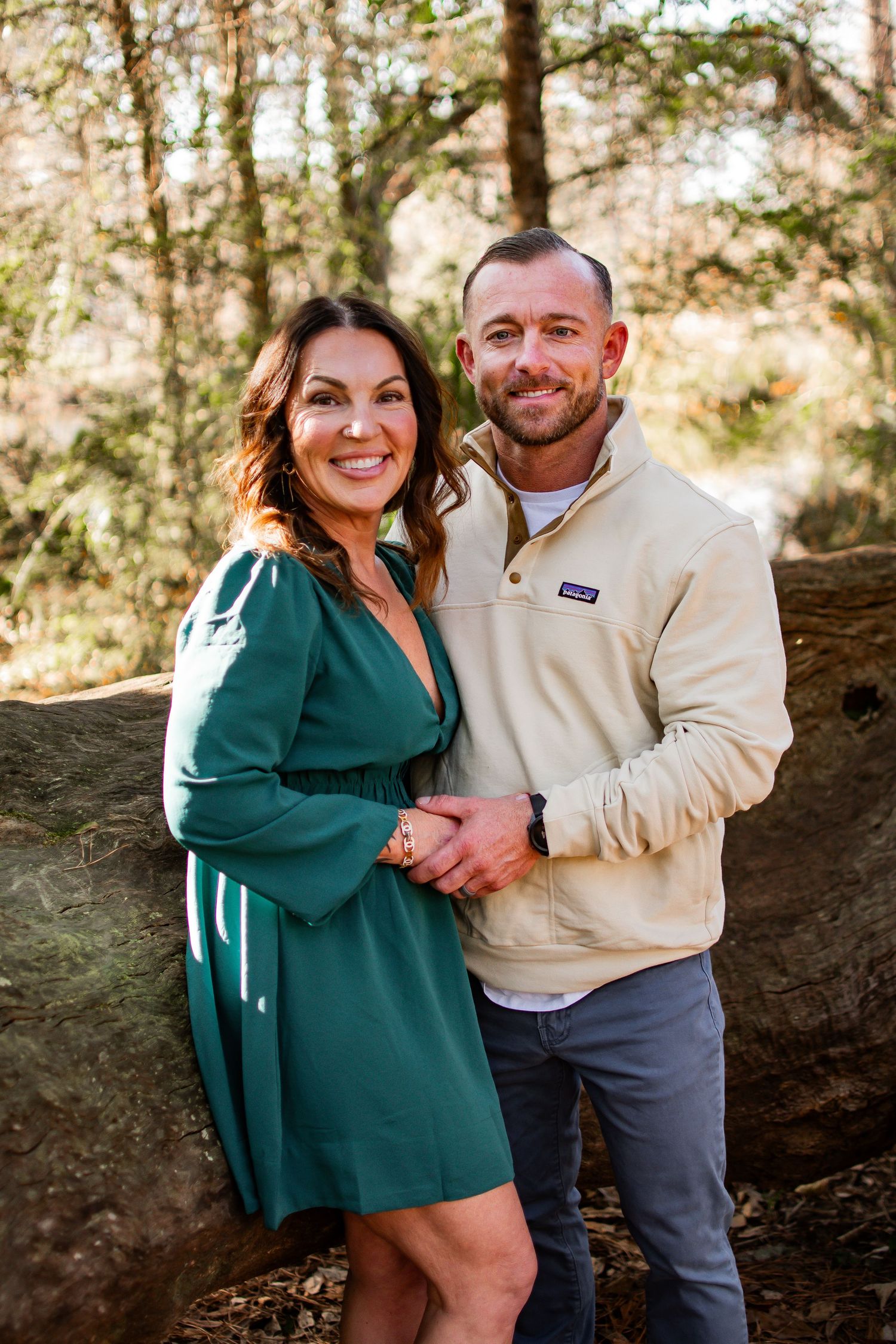 Smiling couple posing together in forest, woman in green dress and man in cream pullover.