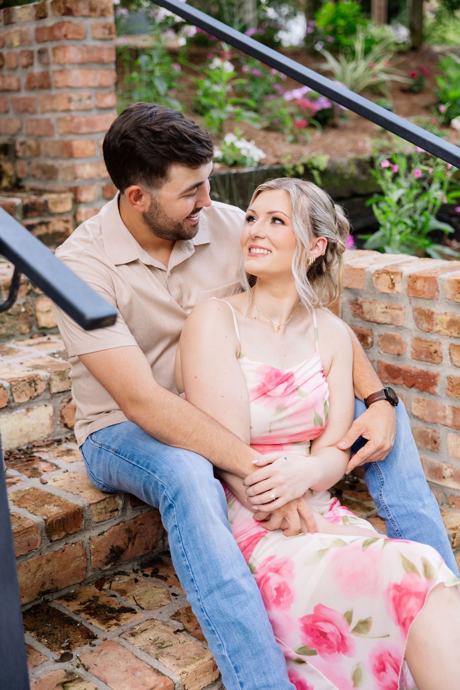 A loving couple sits together on brick steps, gazing at each other, surrounded by colorful garden flowers.