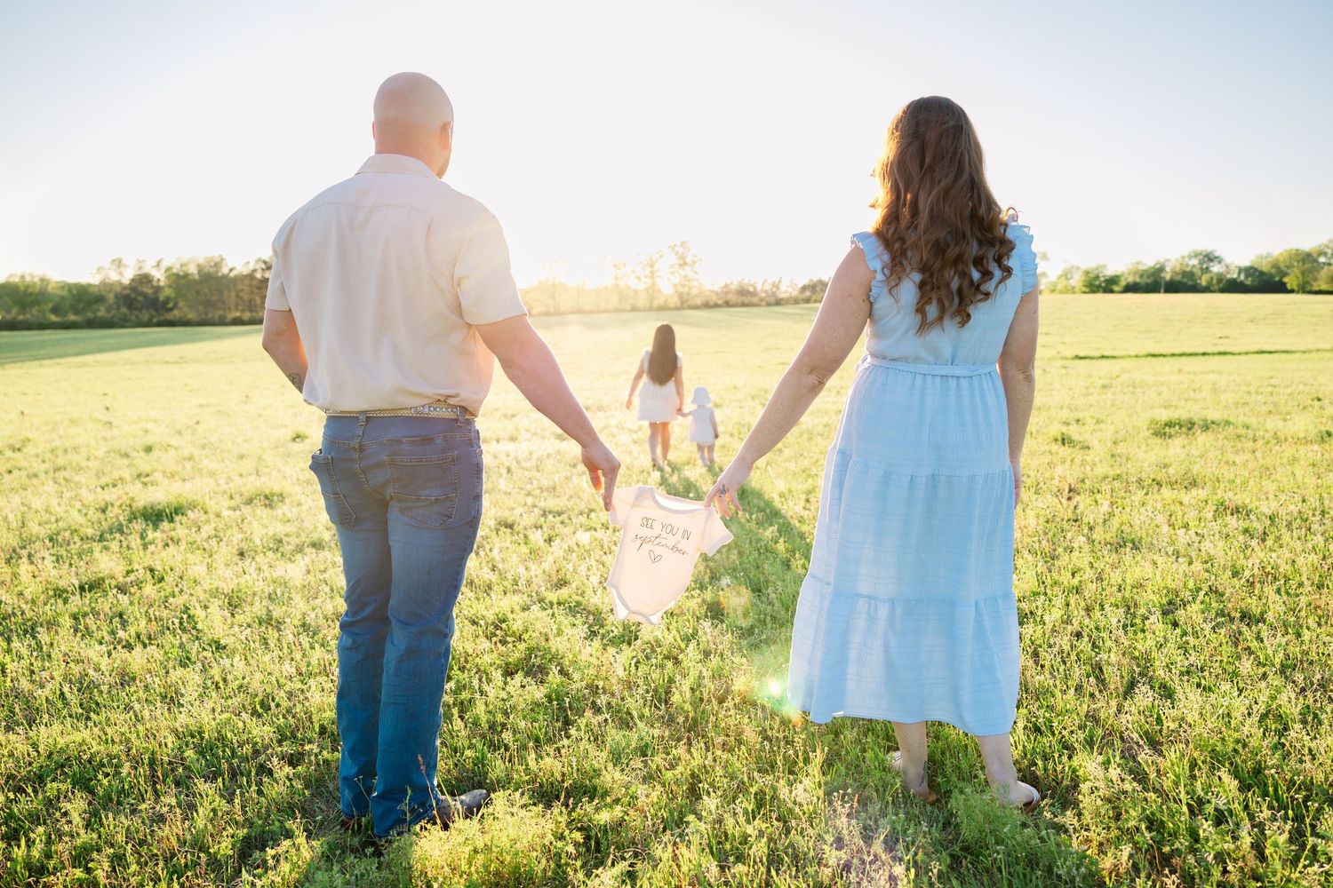 Couple holding hands while daughter runs ahead through lush green field at golden hour.