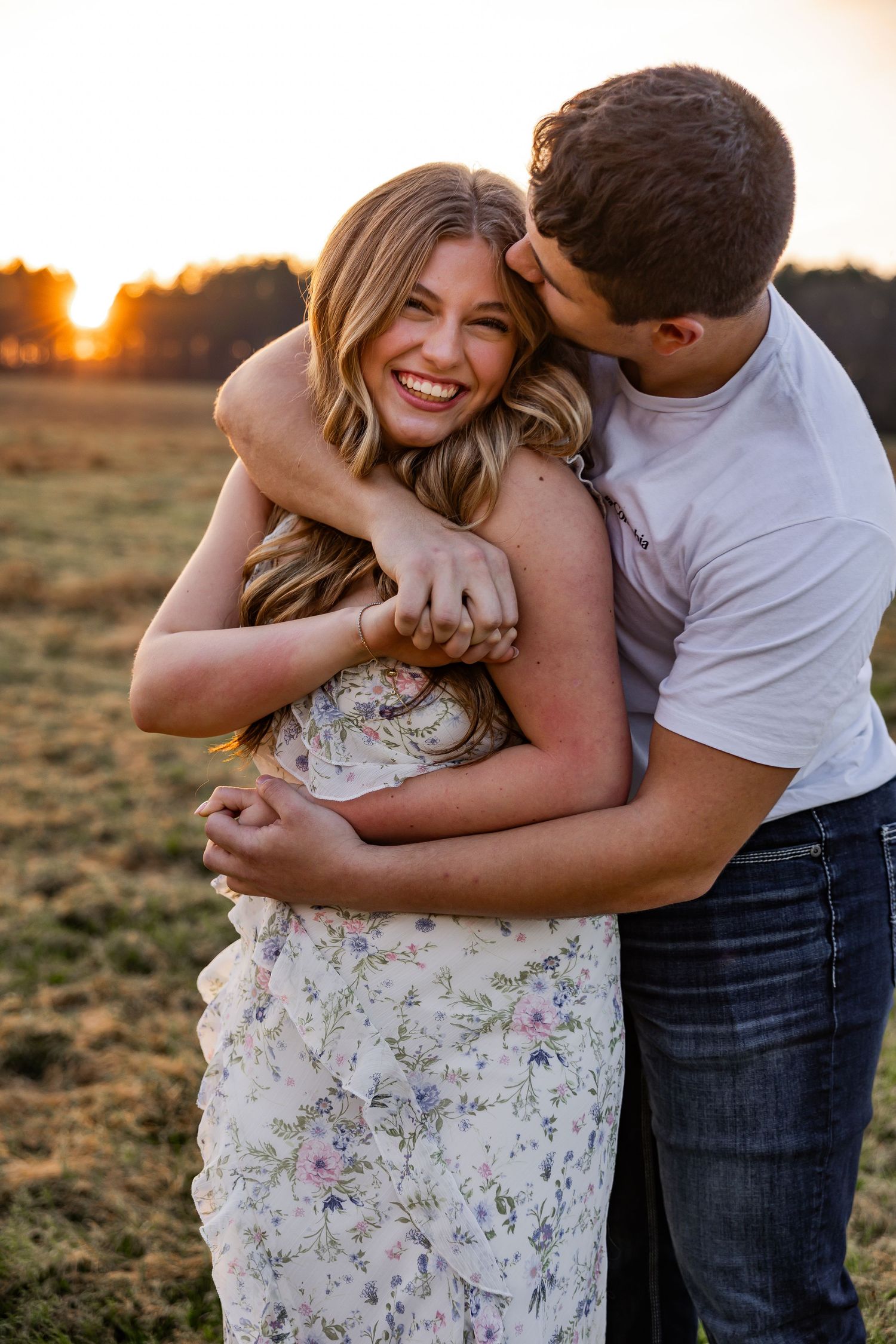 Man hugging woman from behind and kissing her cheek at sunset in an open field, both laughing joyfully.