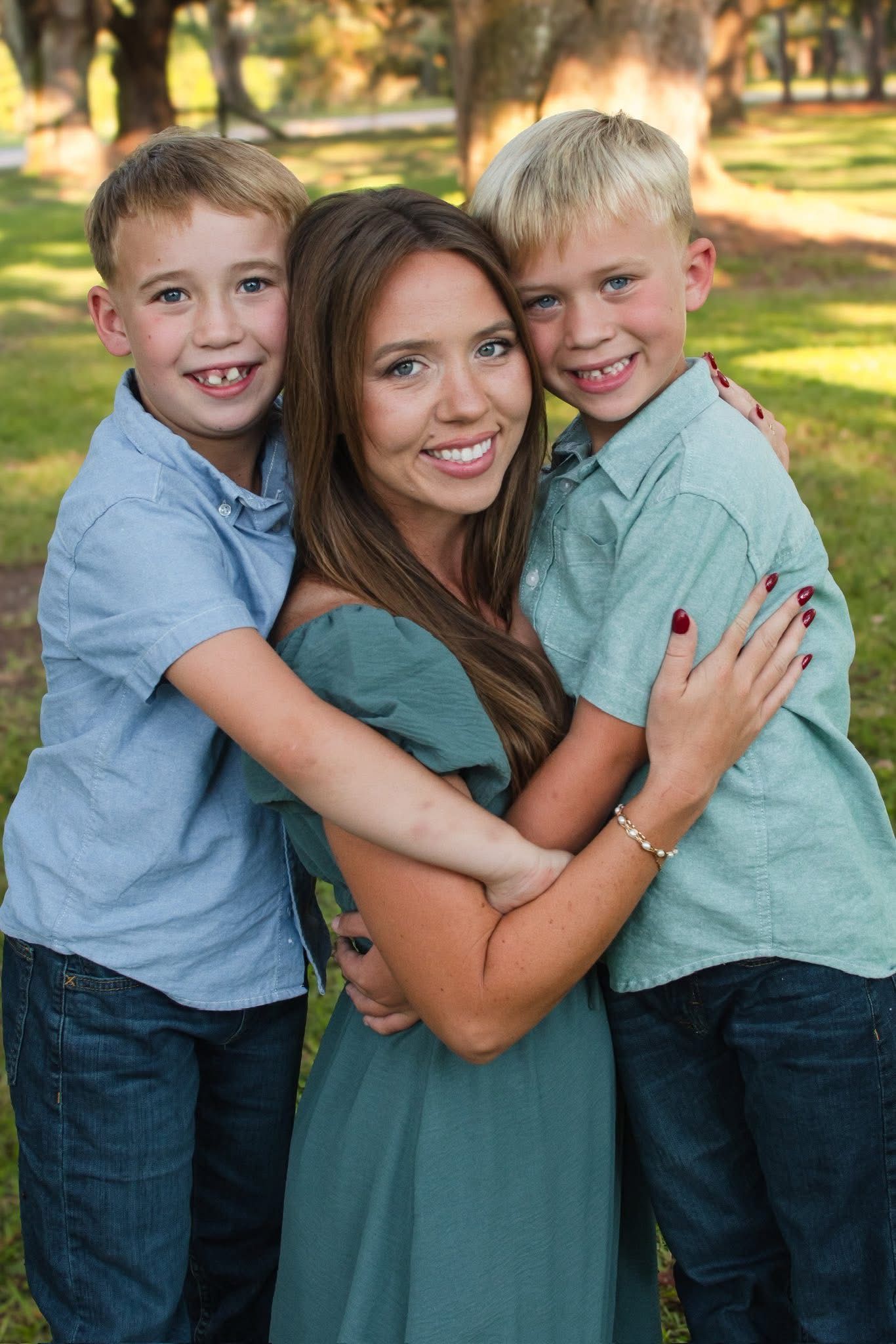 Smiling brunette mother in teal dress embraced by two blonde boys during outdoor family portrait.