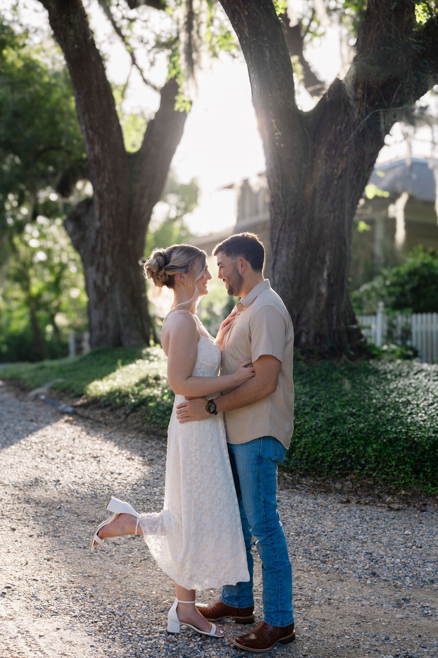 Couple embracing on a sunlit gravel path surrounded by large oak trees, woman in white dress, man in beige shirt and jeans.