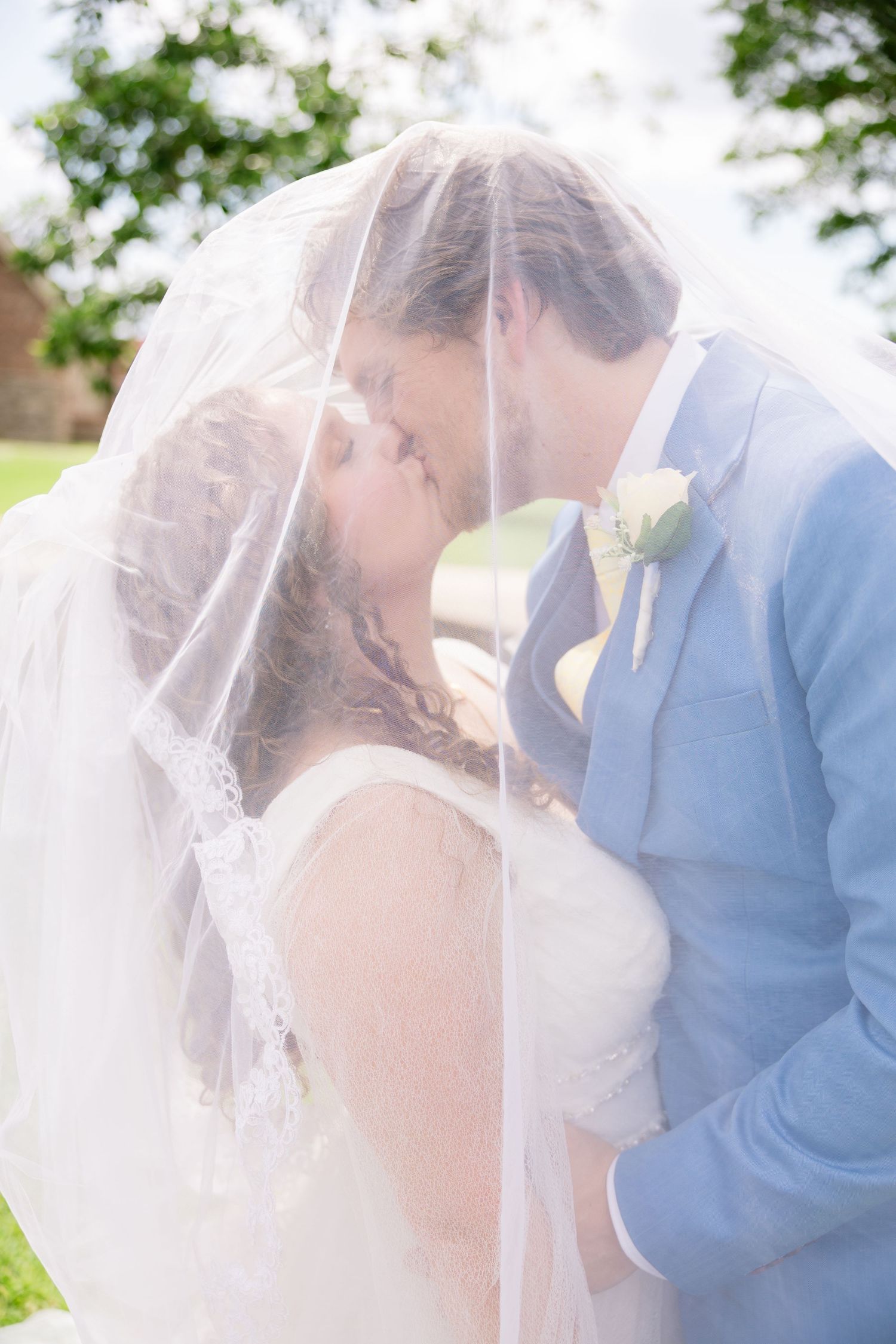 Bride and groom sharing a romantic kiss under a flowing white wedding veil outdoors on their wedding day.