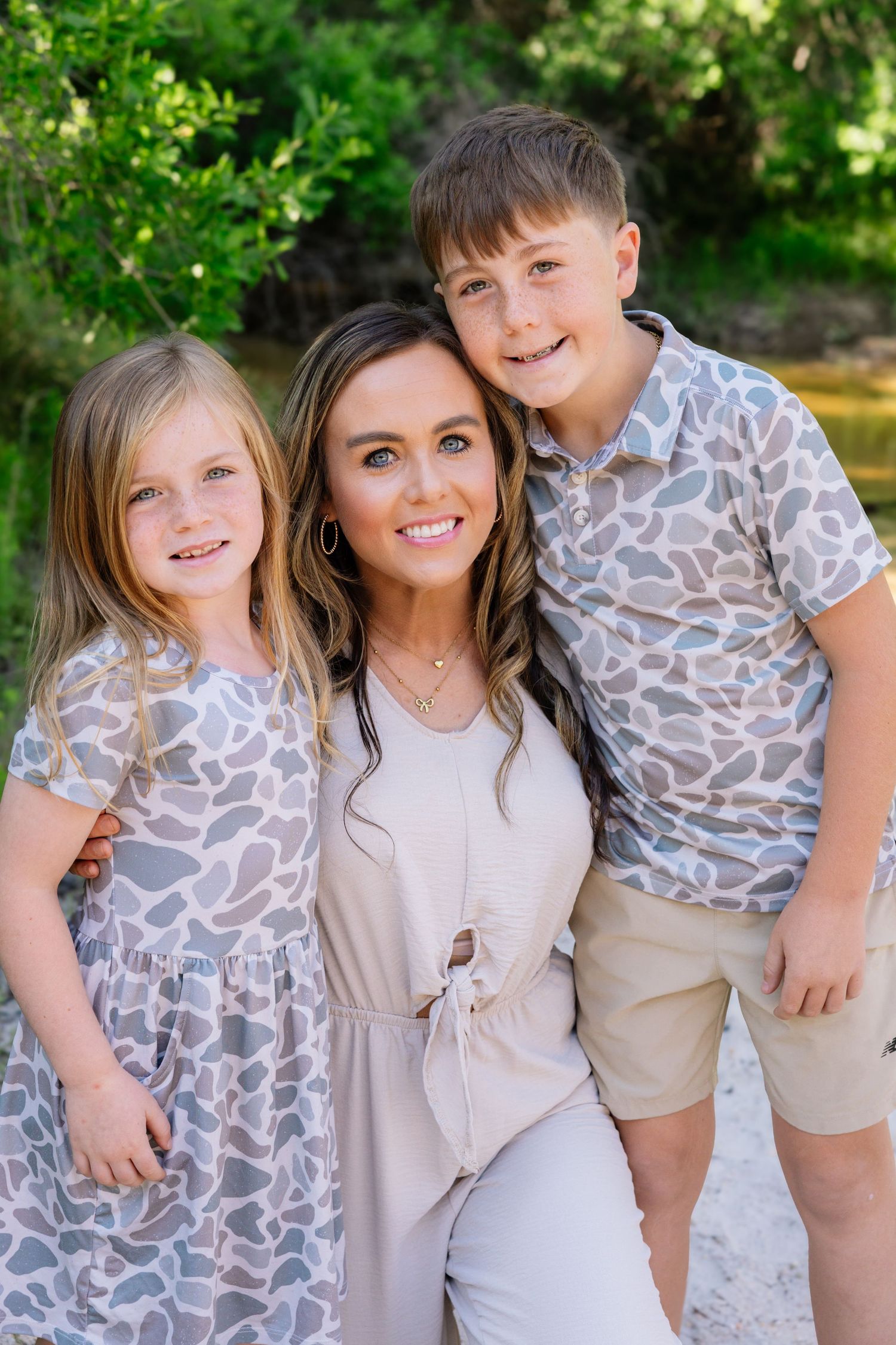 Smiling brunette woman posing outdoors with two young children in matching animal print outfits.