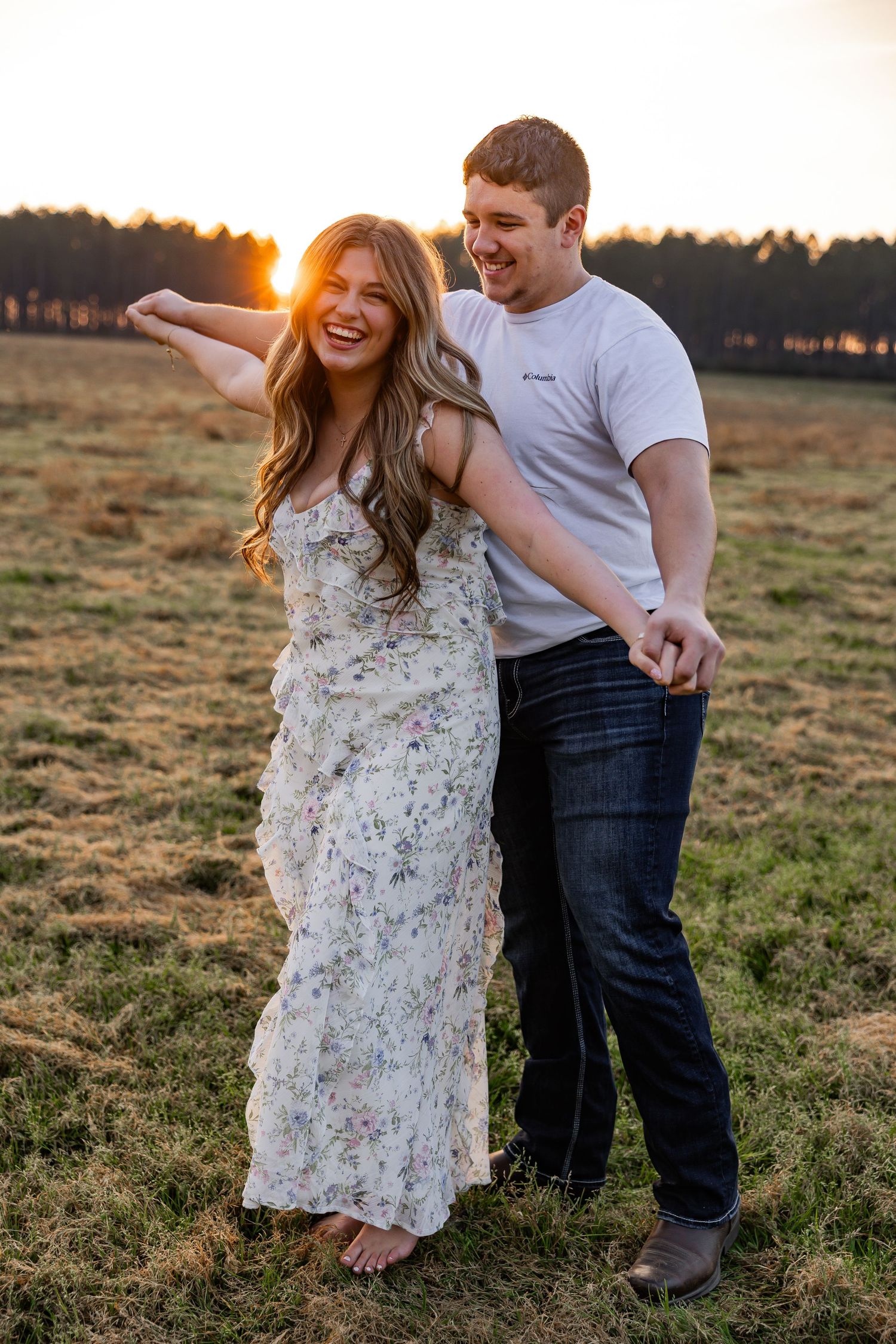 Couple laughing together at golden hour sunset in a field, woman in floral dress and man in white tee.