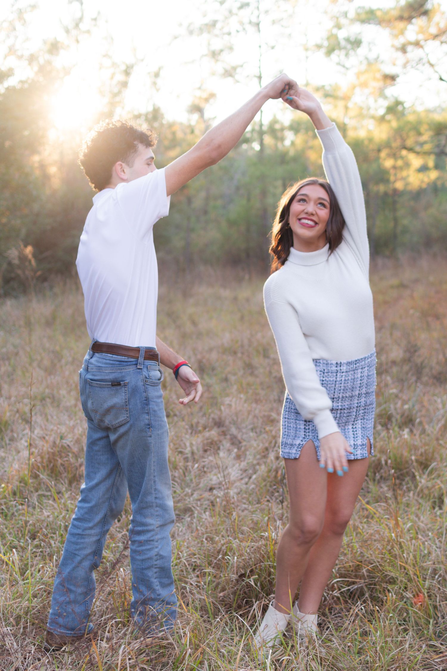 Young man spinning his smiling girlfriend in a sunny open field, both dressed in white with warm golden backlight.
