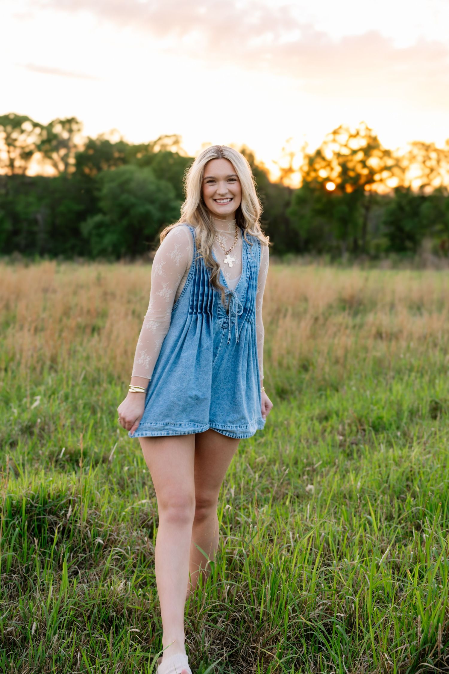 Happy blonde woman in denim romper poses in lush green field during warm sunset.