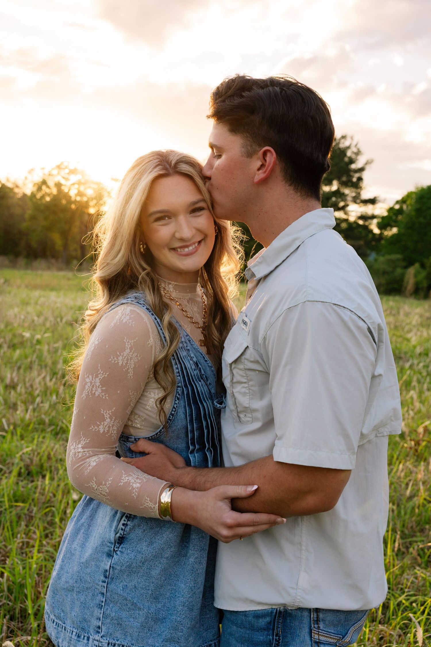Man kisses blonde woman on cheek as she smiles in golden sunset field.